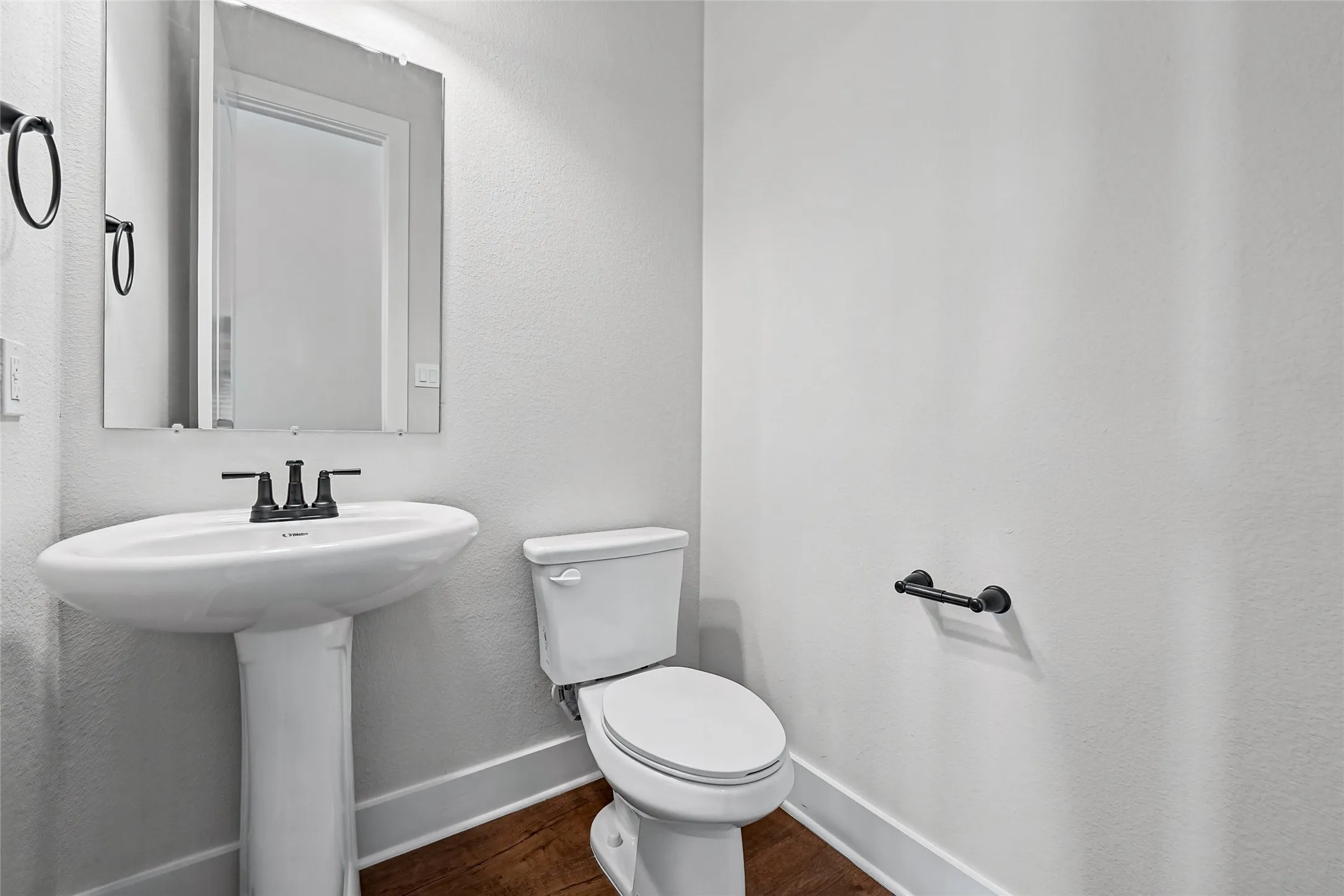Pedestal sink with a black-finish faucet, toilet, wall-mounted mirror, and wood-finish flooring