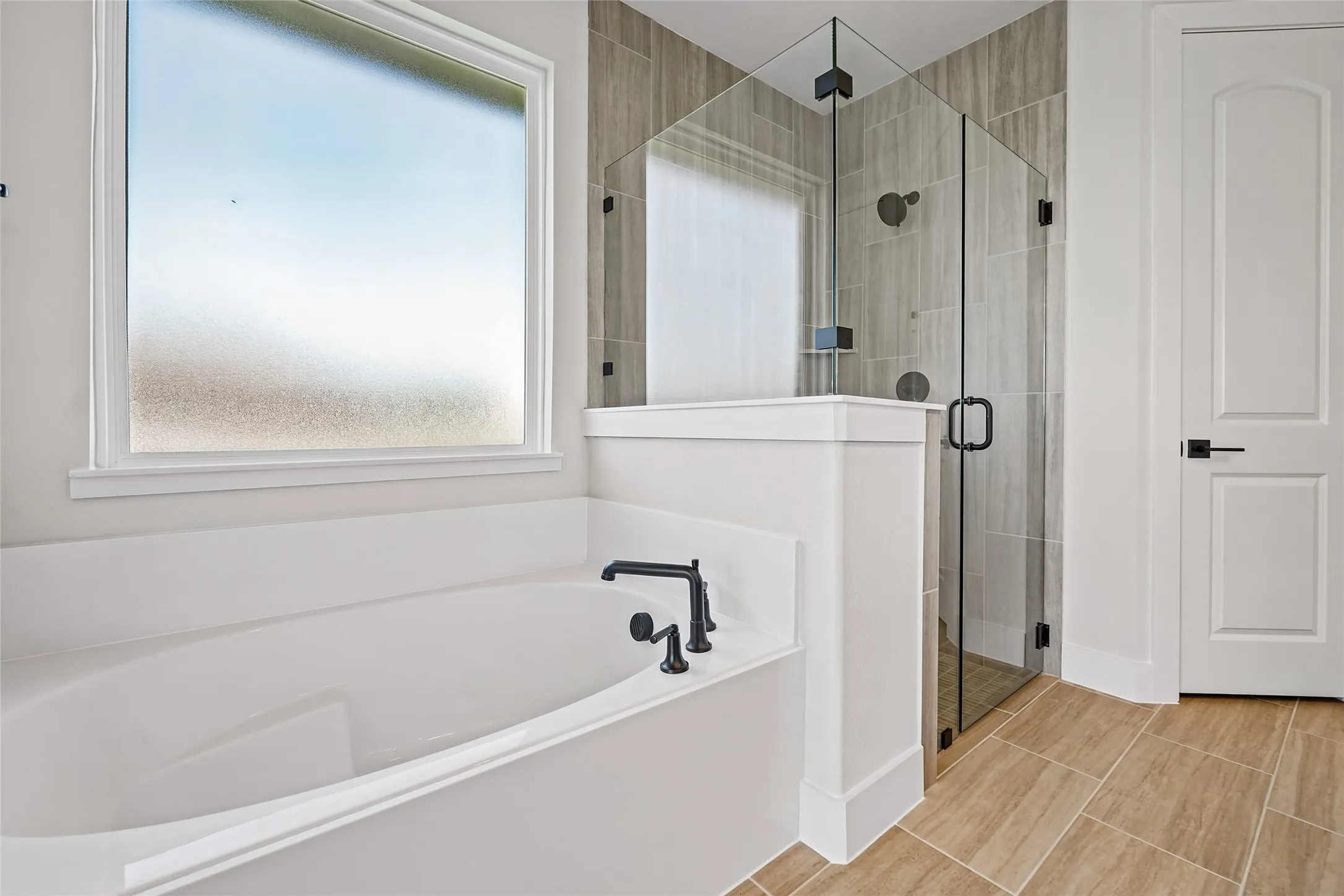 Freestanding soaking tub with matte black fixtures, glass-enclosed shower with tiled walls, and wood-finish tile flooring