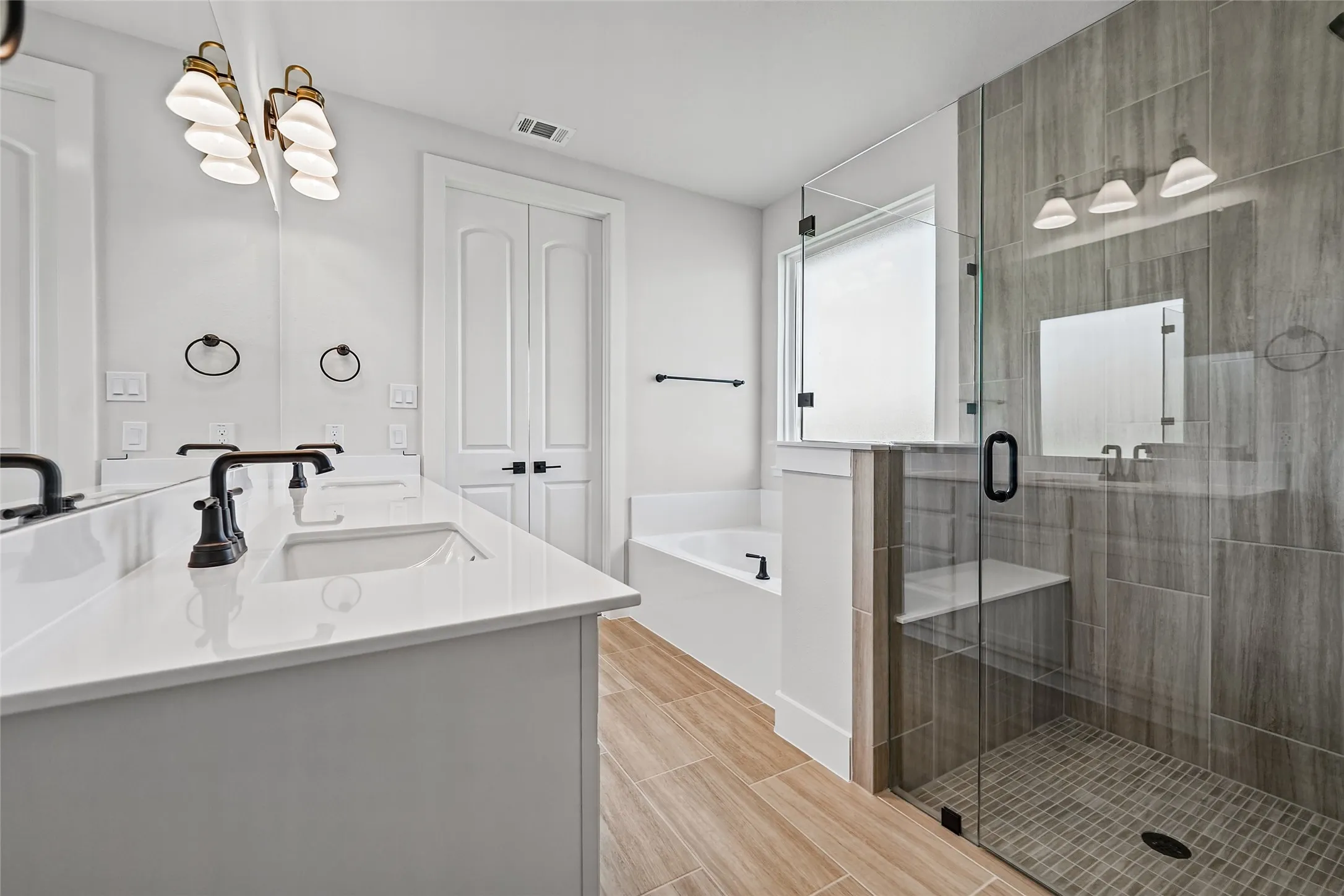 Dual vanity with undermount sinks and dark bronze fixtures, glass-enclosed shower with tile surround and integrated bench, separate soaking tub, and wood-finish flooring