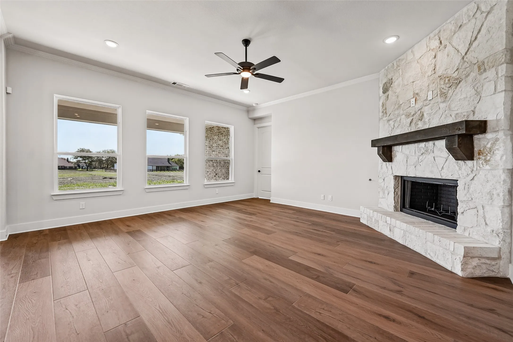Spacious room featuring wood-finish flooring, a stone-clad fireplace with a dark wood mantel, three prominent windows, and a ceiling fan with integrated lighting