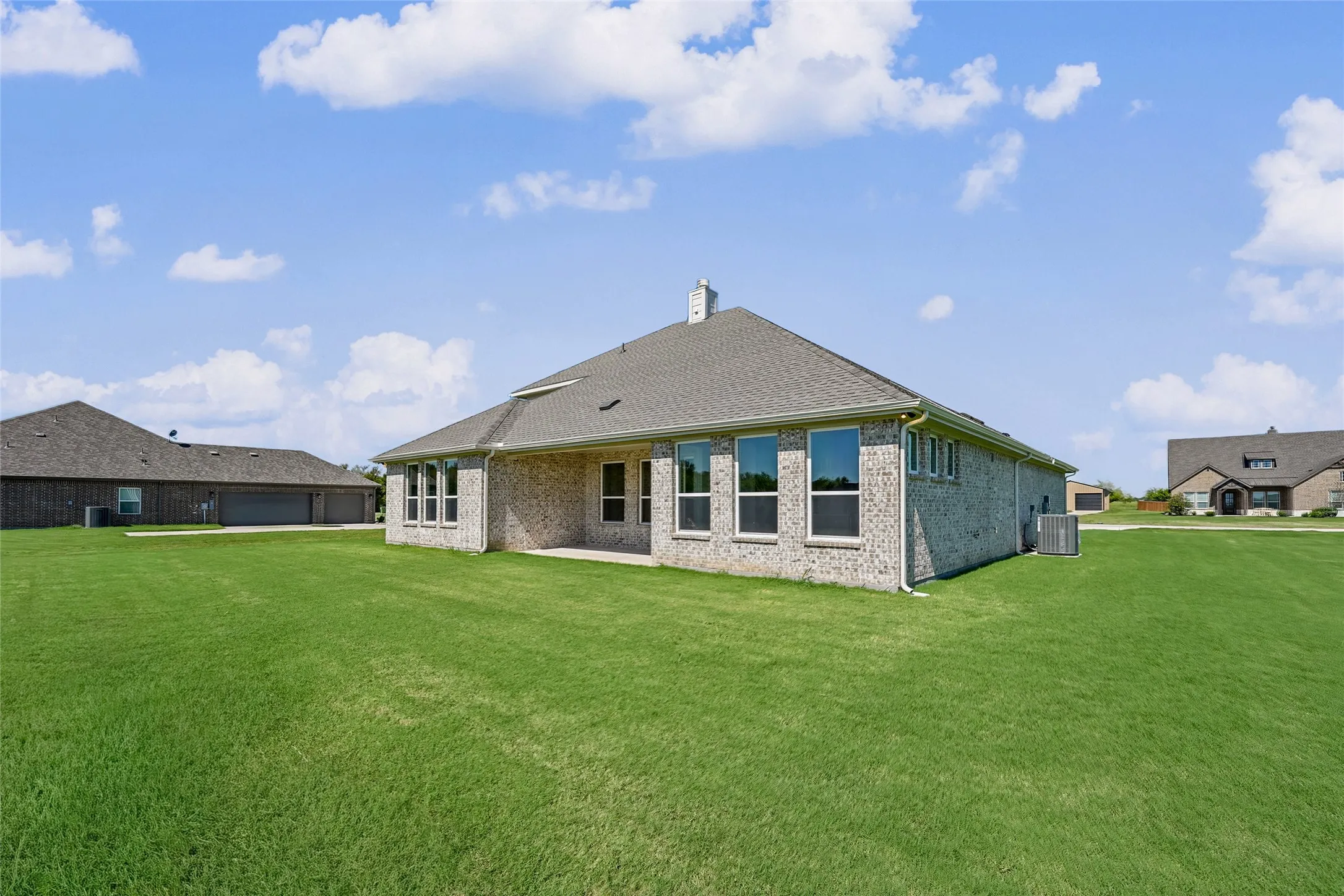 Expansive green lawn surrounds the property, featuring a brick exterior, covered patio, and a shingled roof