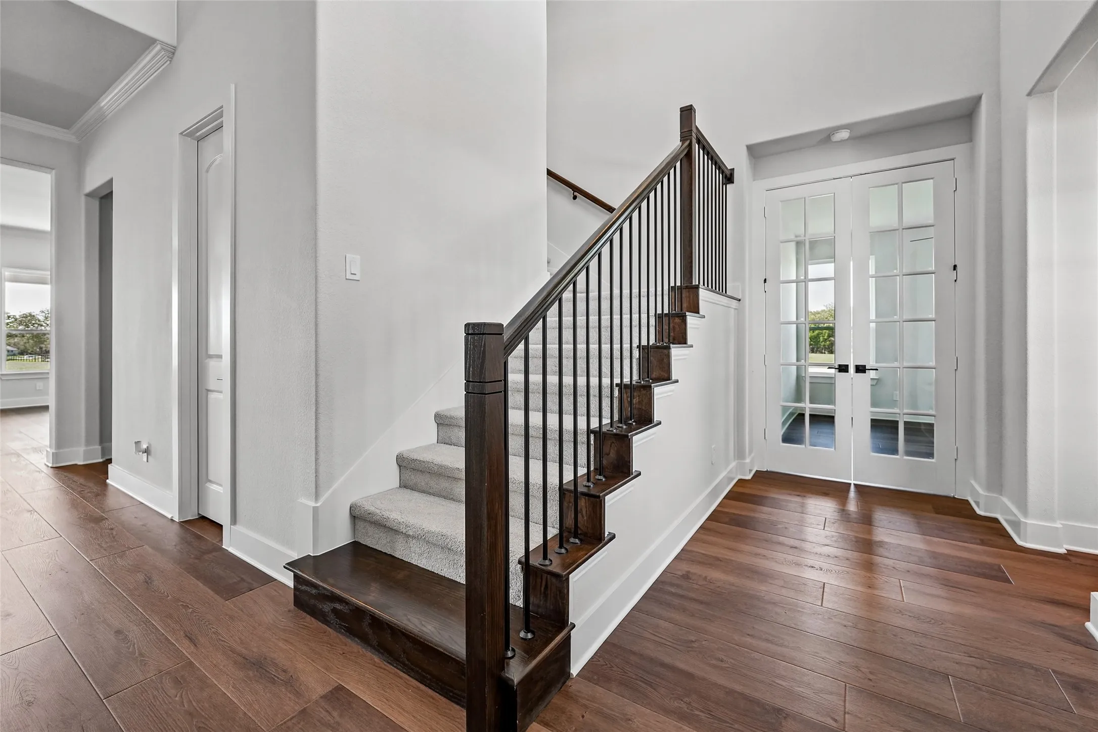 Wood-finish flooring, staircase with dark wood newel posts and wrought iron balusters, light-toned carpeted stair treads, white French doors with divided lites, and crisp white trim throughout