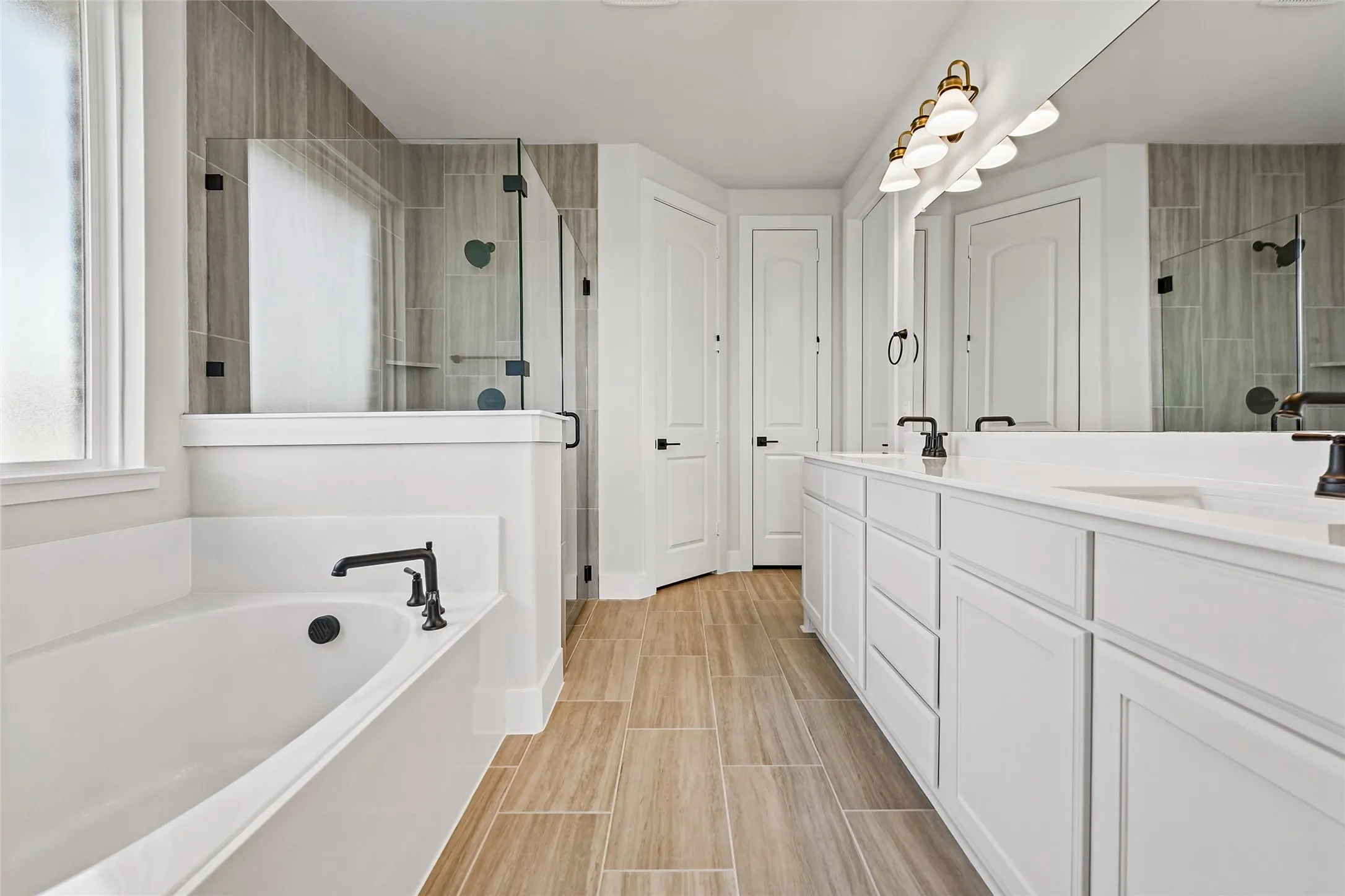 Dual vanity with white cabinetry, built-in soaking tub, walk-in shower with glass enclosure, wood-finish flooring, and wall-mounted lighting