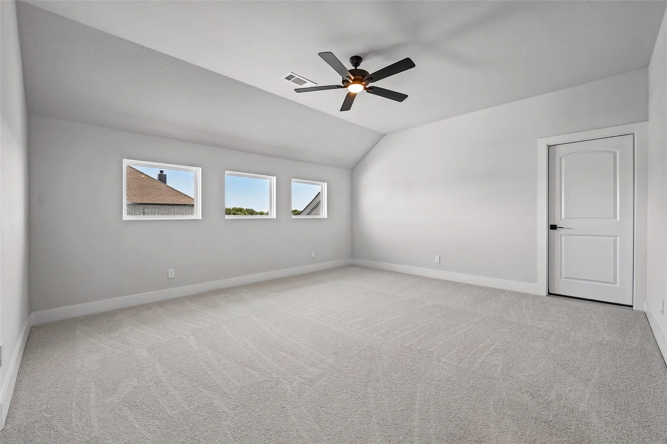 Bright room with carpet flooring, a vaulted ceiling, three transom windows, a ceiling fan with integrated lighting, and a white interior door