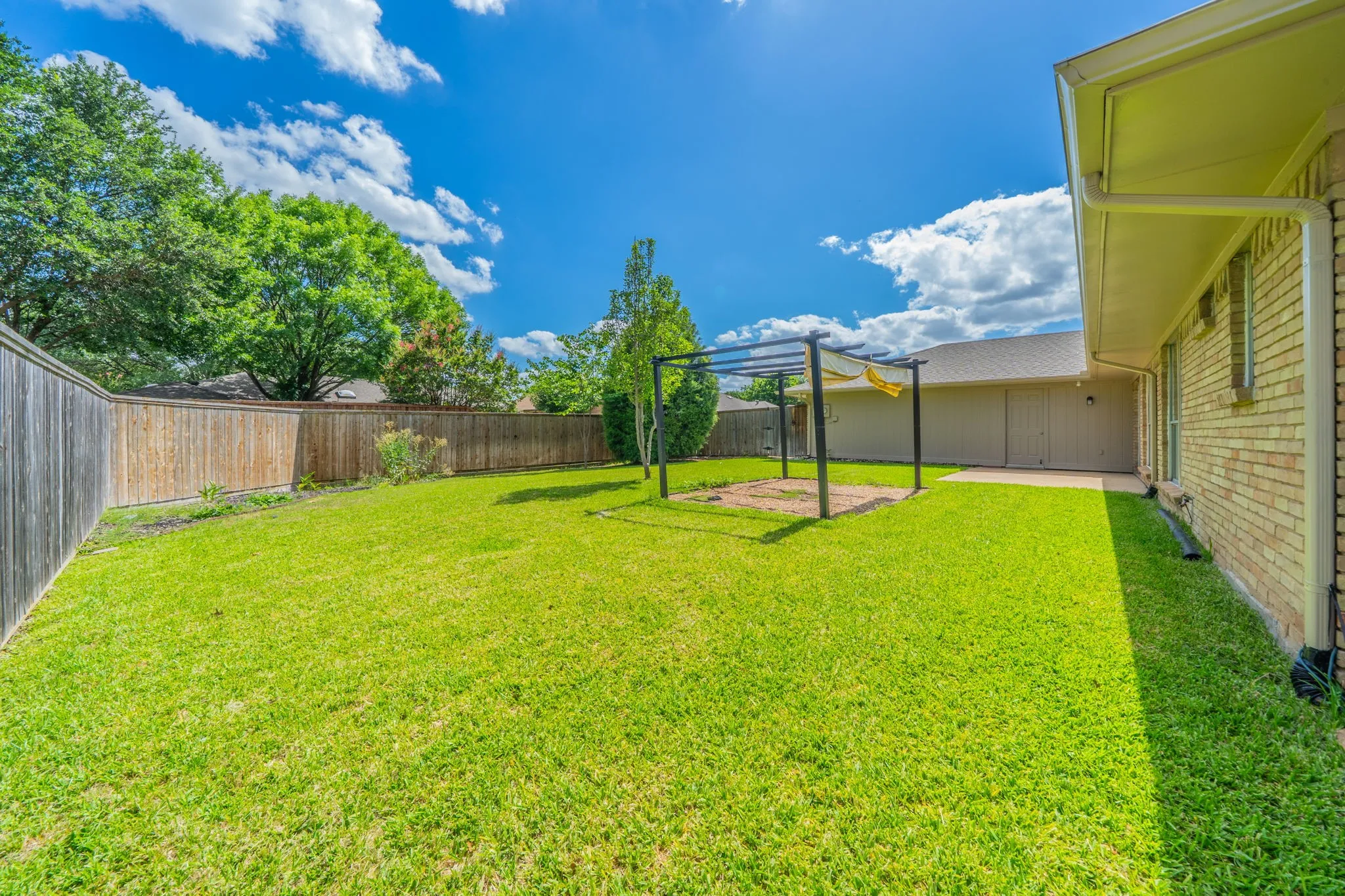 Expansive green lawn featuring a privacy fence, a metal pergola, mature perimeter trees, and a brick exterior with a concrete patio