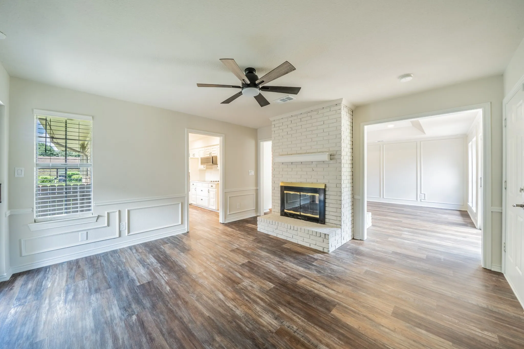 Living area featuring wood-finish flooring, a white-painted brick fireplace with a black firebox, a ceiling fan, and wainscoting with decorative trim