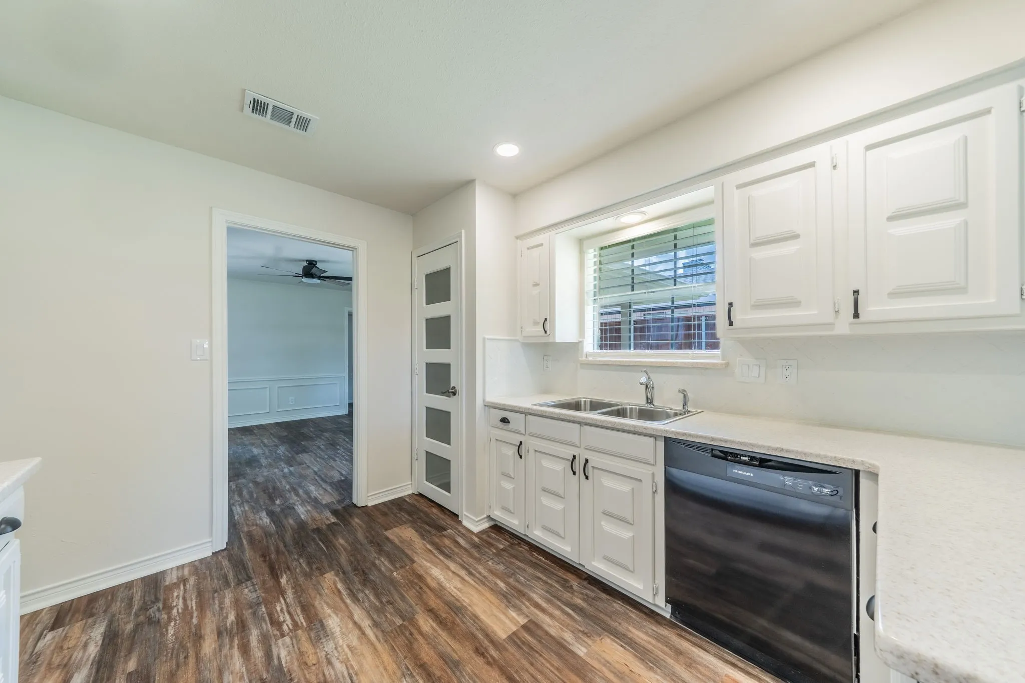 Kitchen featuring wood-finish flooring, white cabinetry, a double basin sink, a black built-in dishwasher, and a window with horizontal blinds