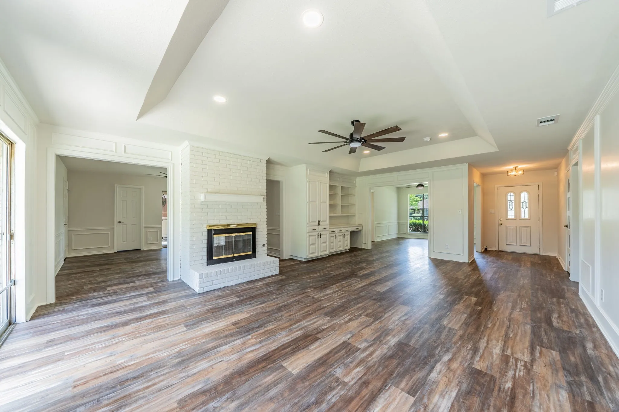 Spacious open concept living area featuring wood-finish flooring, a white painted brick fireplace with a brass-trimmed insert, and a tray ceiling with a ceiling fan