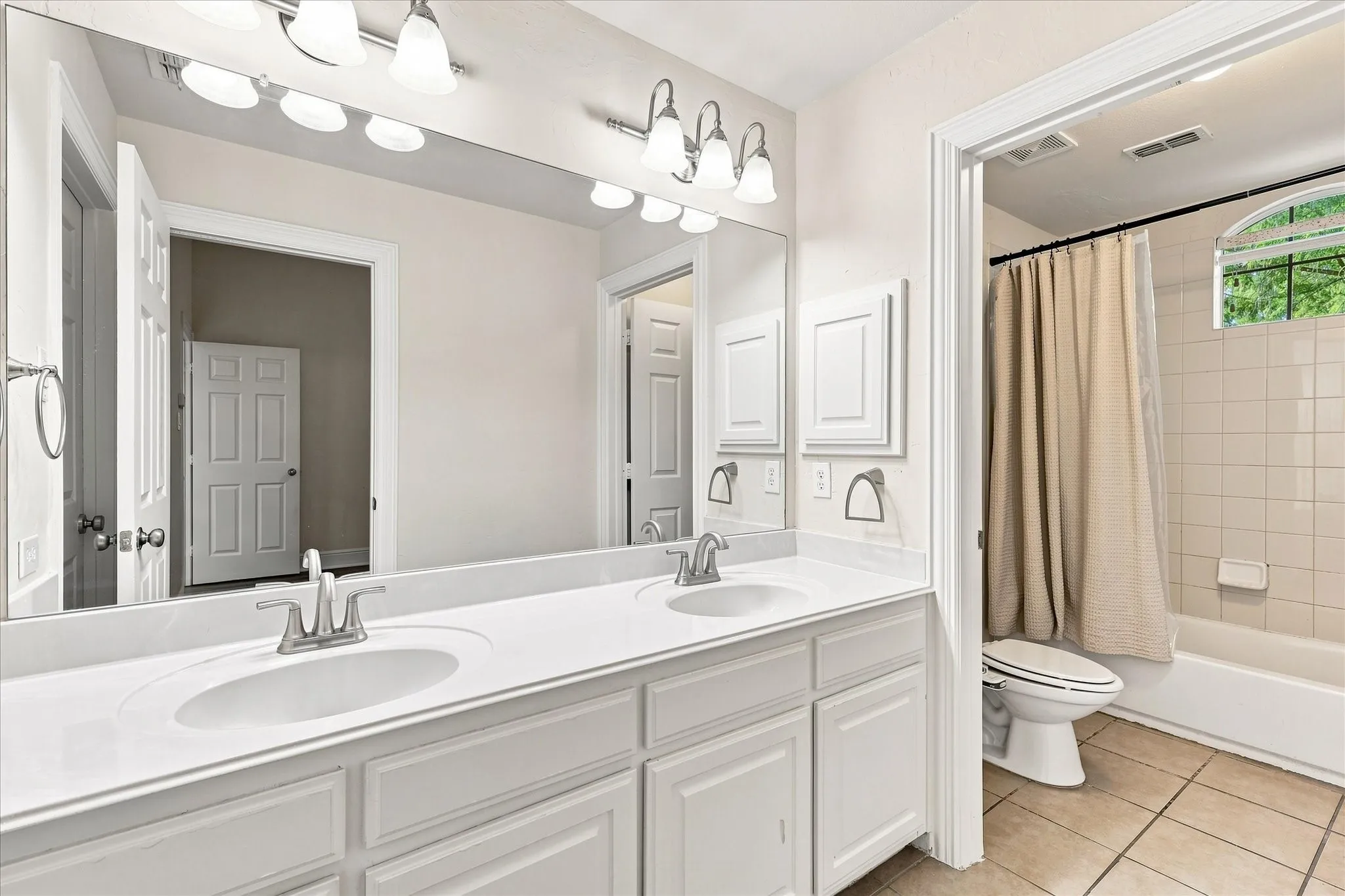 Dual vanity with integrated sinks and chrome fixtures, white tile flooring, and a tiled shower-tub combination with an arched window
