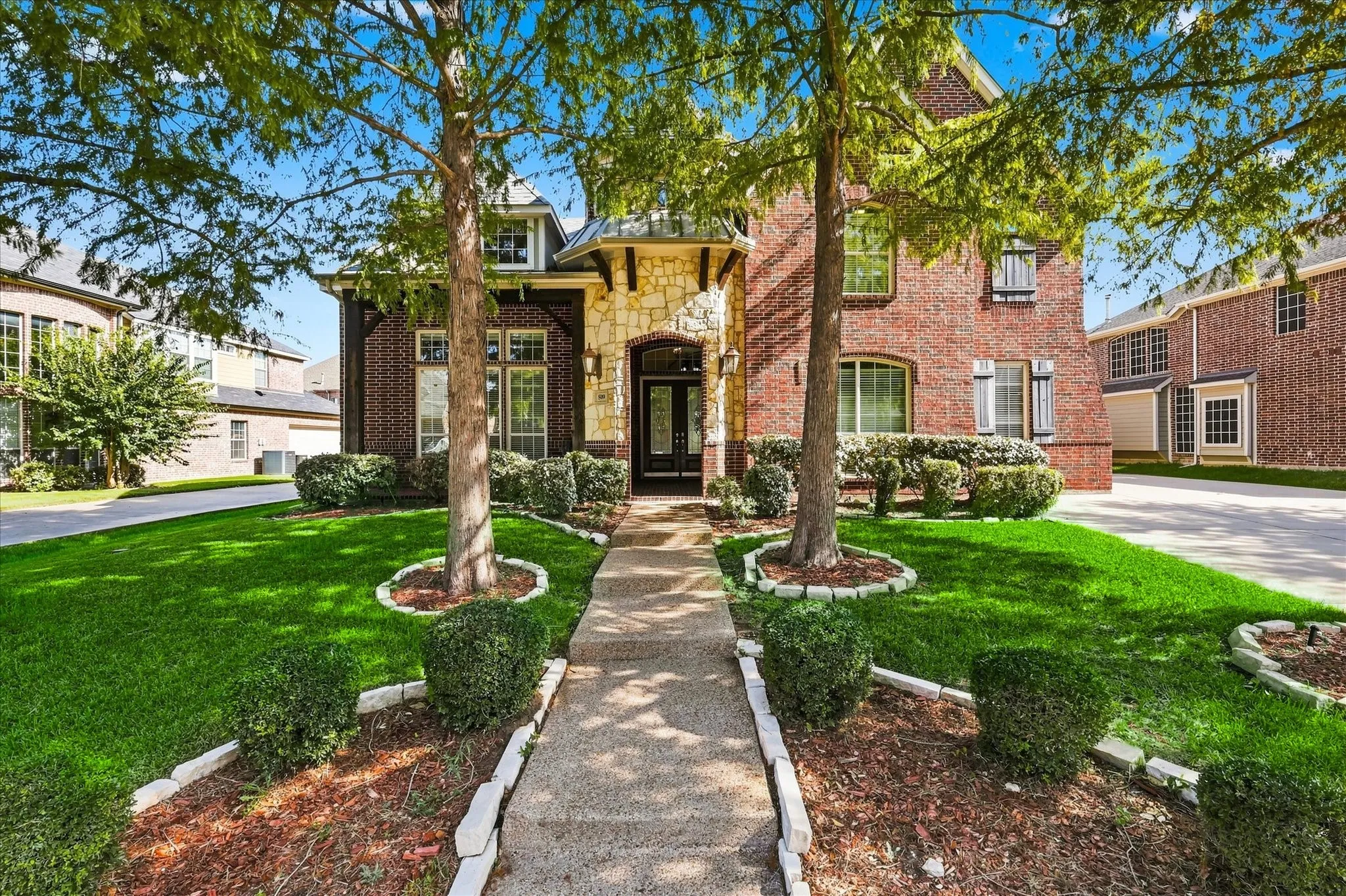 Brick and stone facade, arched entryway, gabled roofline, mature trees, concrete pathway