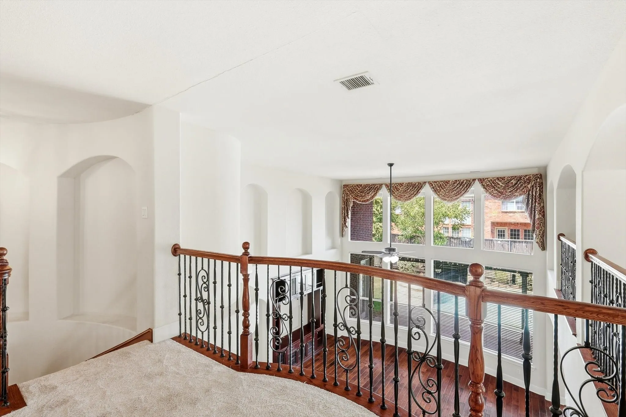 Second-story landing featuring curved wood and wrought iron balustrade, recessed wall niches, and a full-height window array with decorative valances