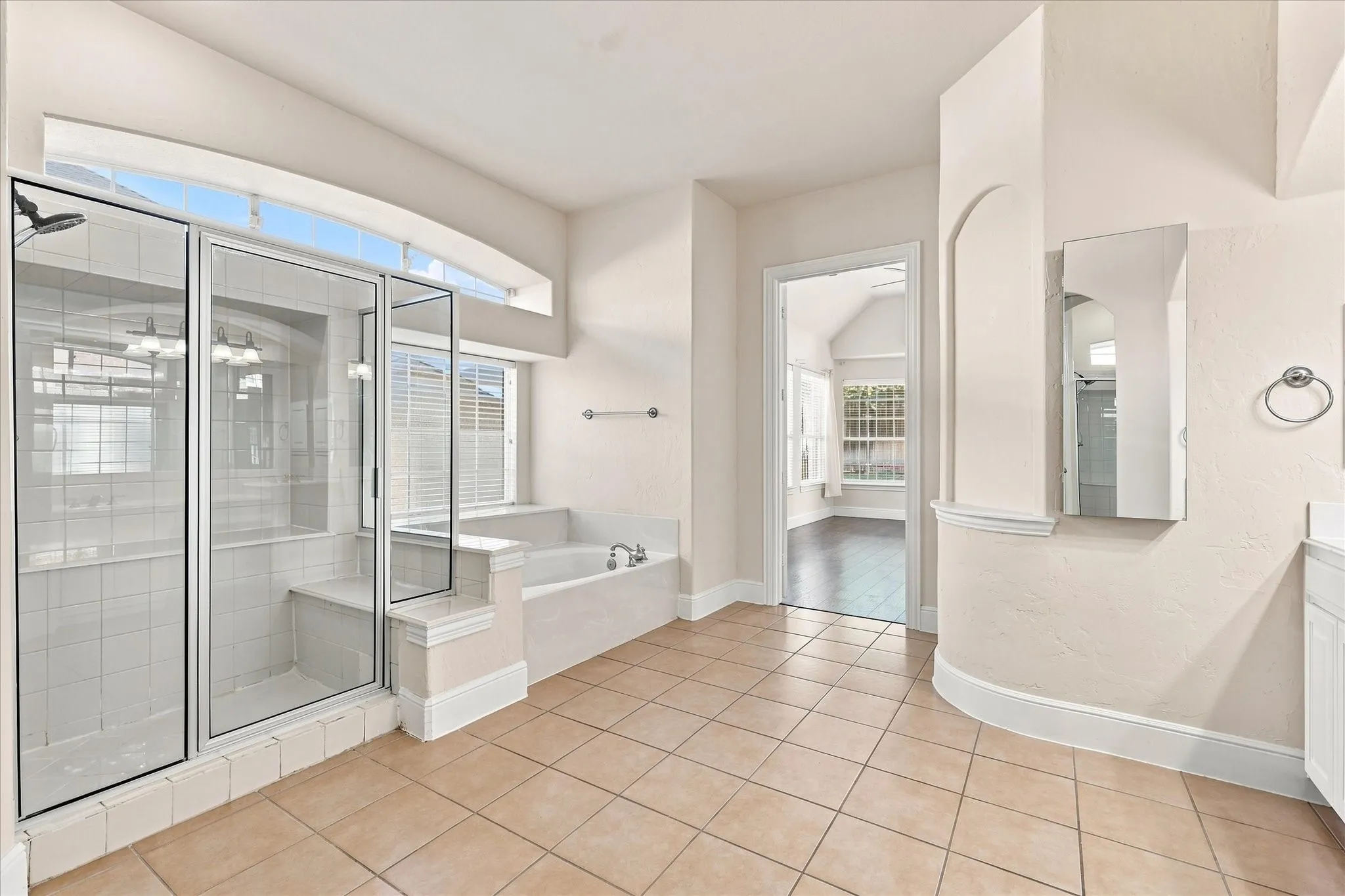 Spa-inspired primary bathroom featuring a frameless glass-enclosed shower with tile surround, soaking tub with deck-mounted fixtures, dual vanity with medicine cabinet, tile flooring, and abundant natural light from transom windows