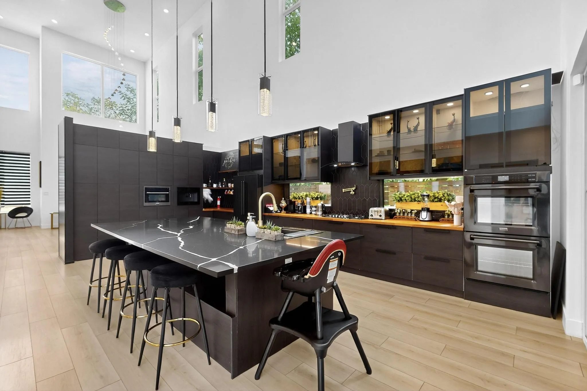 Kitchen featuring a large central island with a waterfall countertop, integrated dual ovens, glass-front upper cabinetry, wood-finish flooring, and high ceilings with upper windows