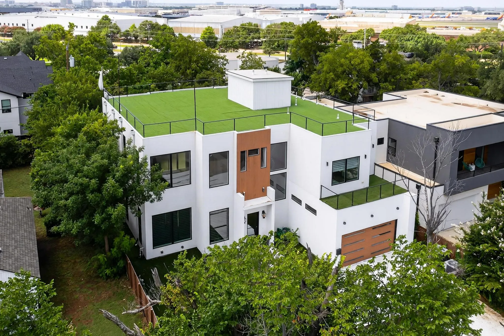 Modern multi-story residence featuring a white stucco exterior with wood-tone accent panels, a wood-finish garage door, black-framed windows, and multiple rooftop areas with artificial turf and black metal railings