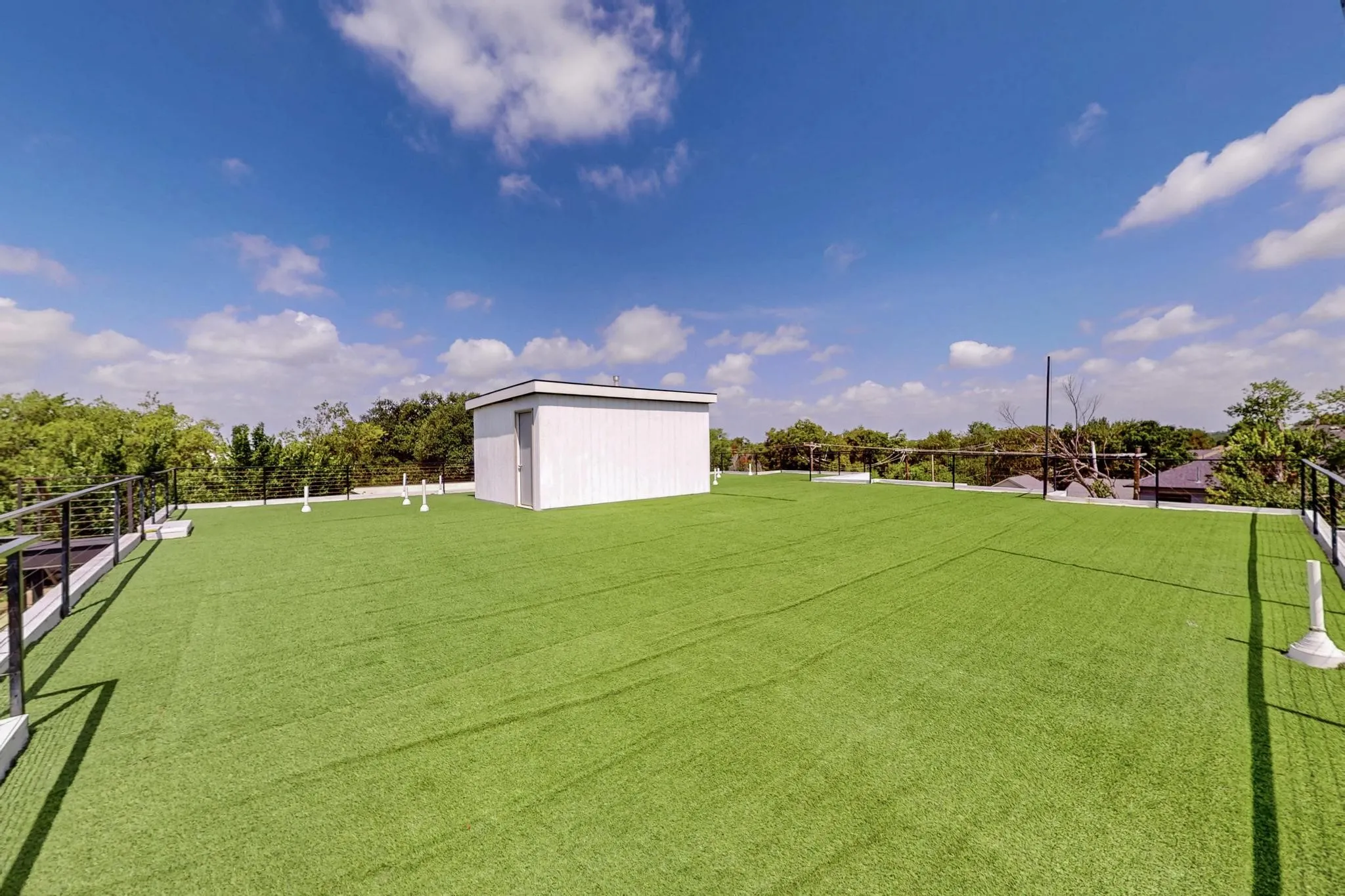 Expansive rooftop terrace featuring artificial turf, black metal railings, a white utility structure, and surrounding tree canopy