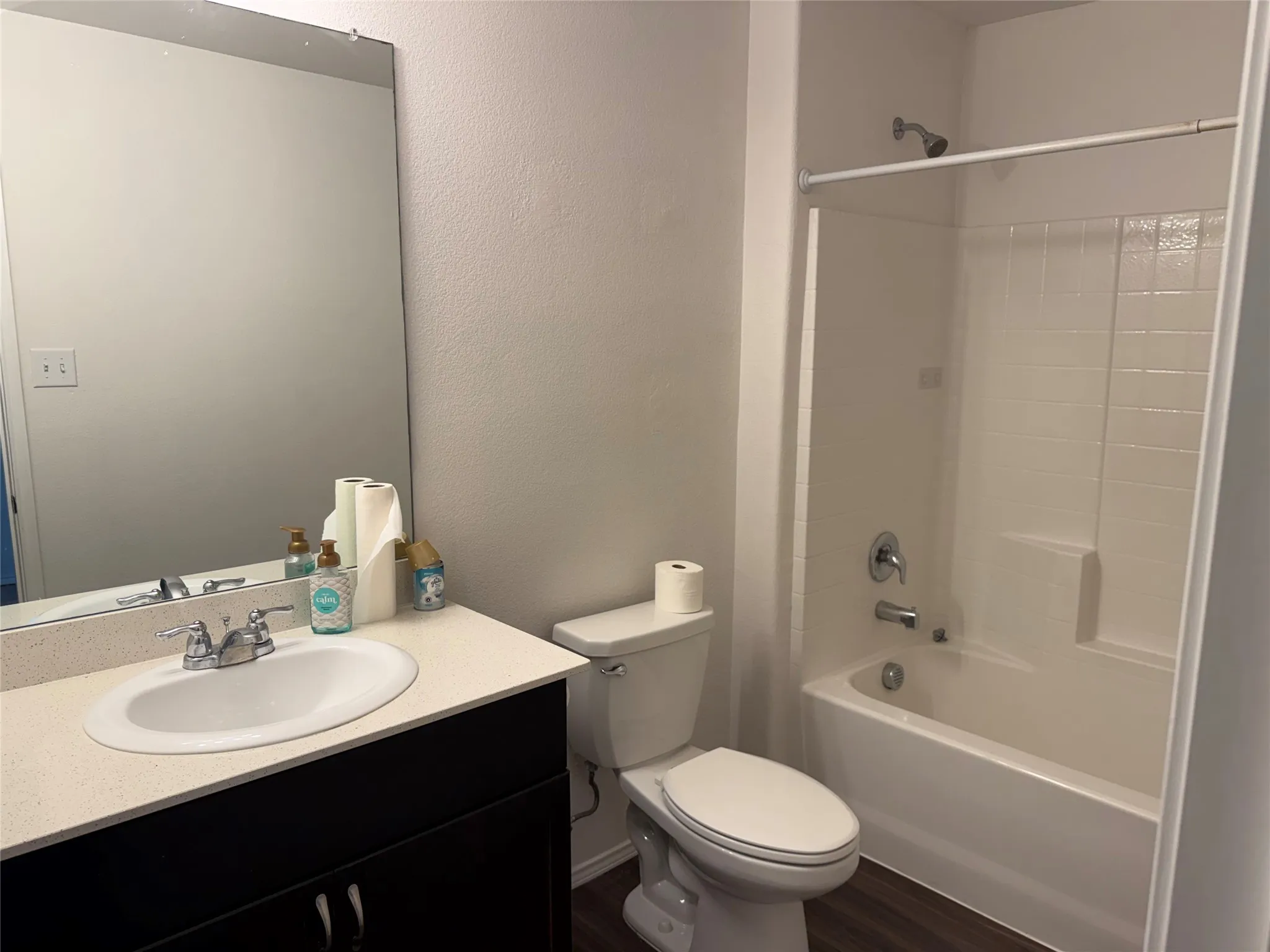 Contemporary bathroom featuring a dark wood-finish vanity with a light-toned countertop, an integrated sink with a chrome faucet, a standard toilet, and a white tub/shower combination with a textured surround