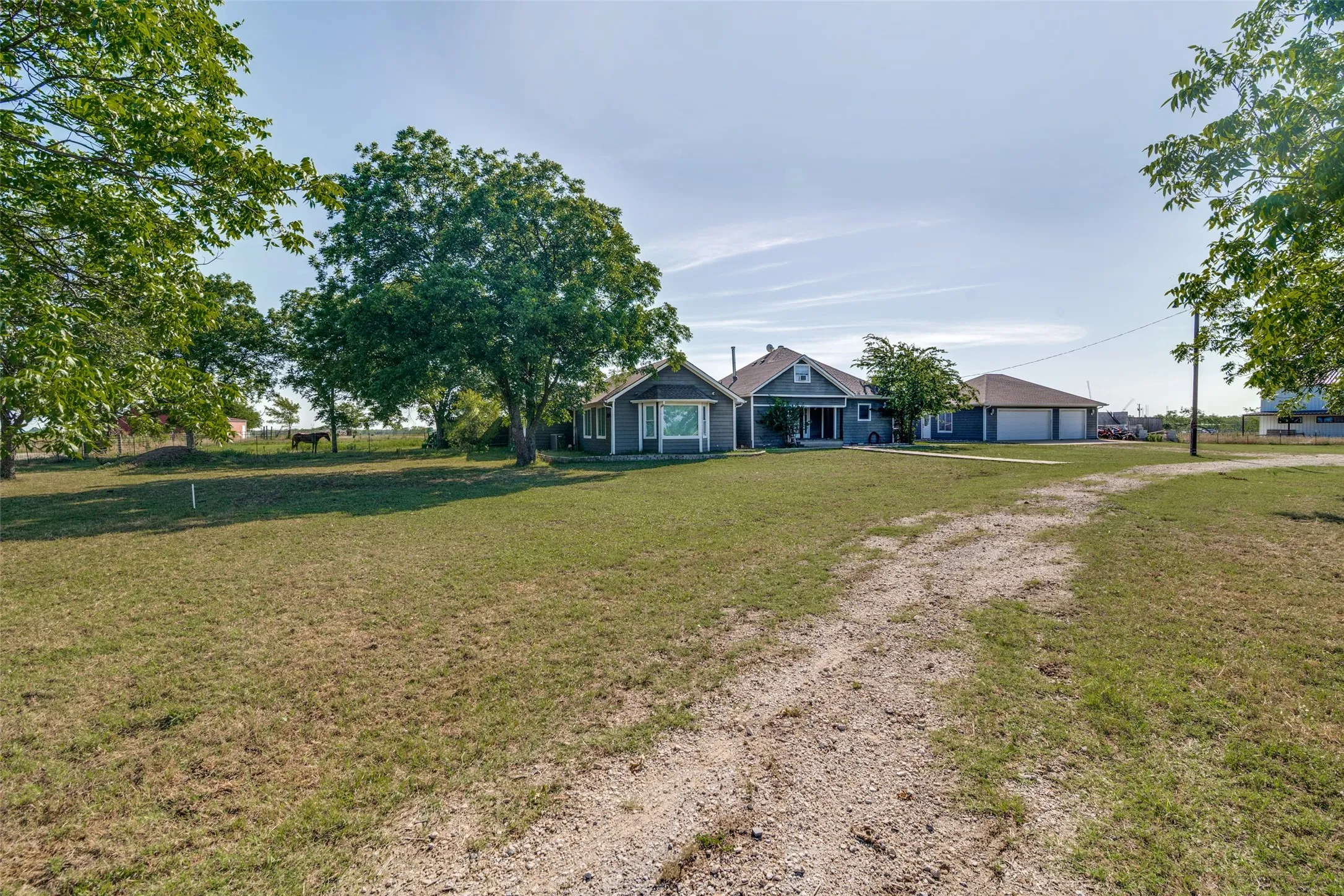 Spacious property featuring a single-story residence with a detached garage, expansive grassy areas, mature trees, and a gravel driveway