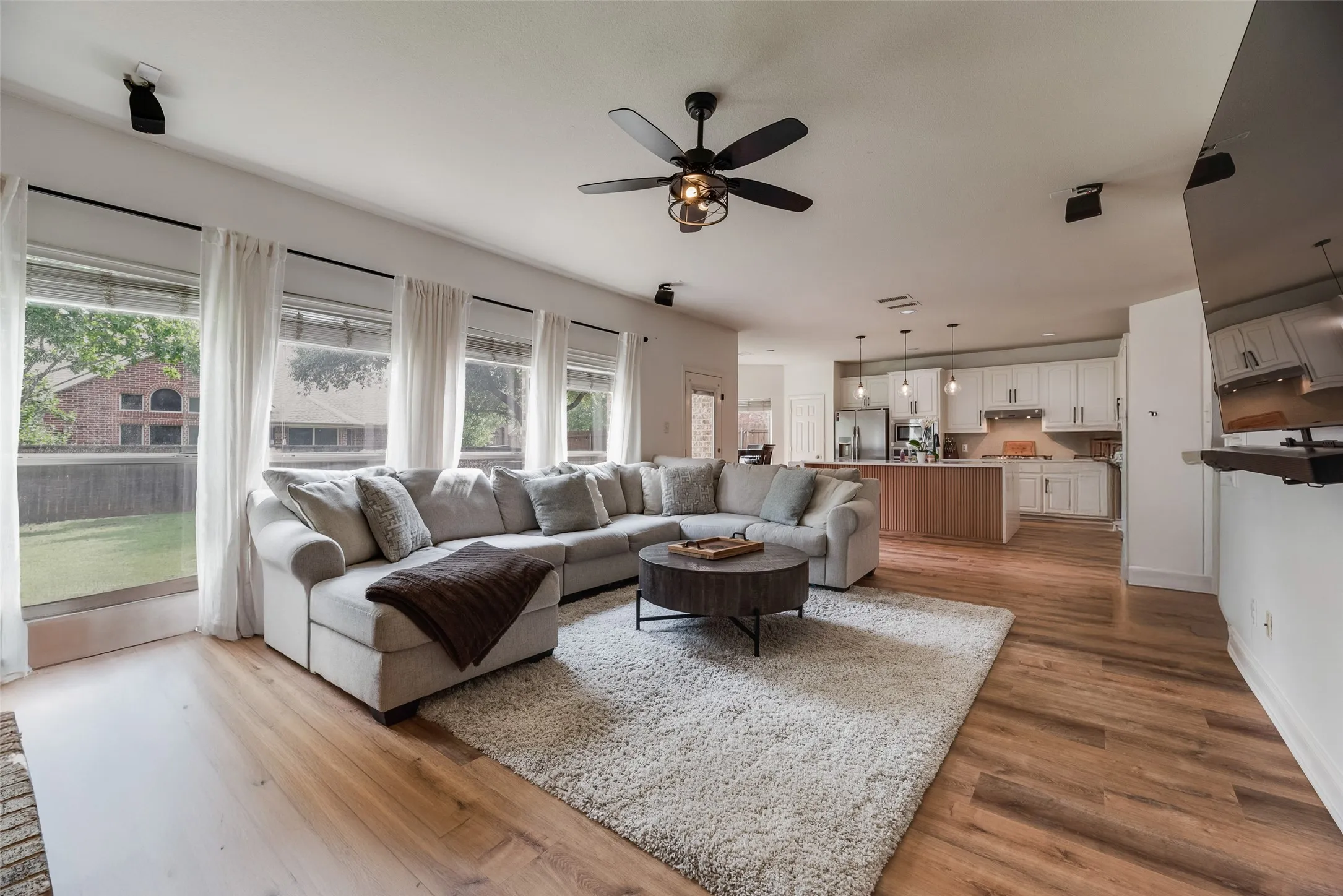 Open-concept living area with wood-finish flooring, a contemporary ceiling fan, and multiple large windows overlooking the backyard