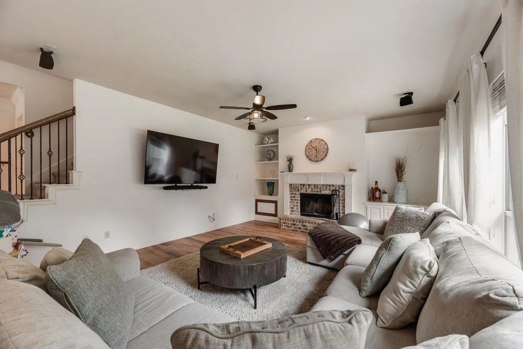 Spacious living area featuring wood-finish flooring, a brick fireplace with a mantle, built-in shelving, and a ceiling fan