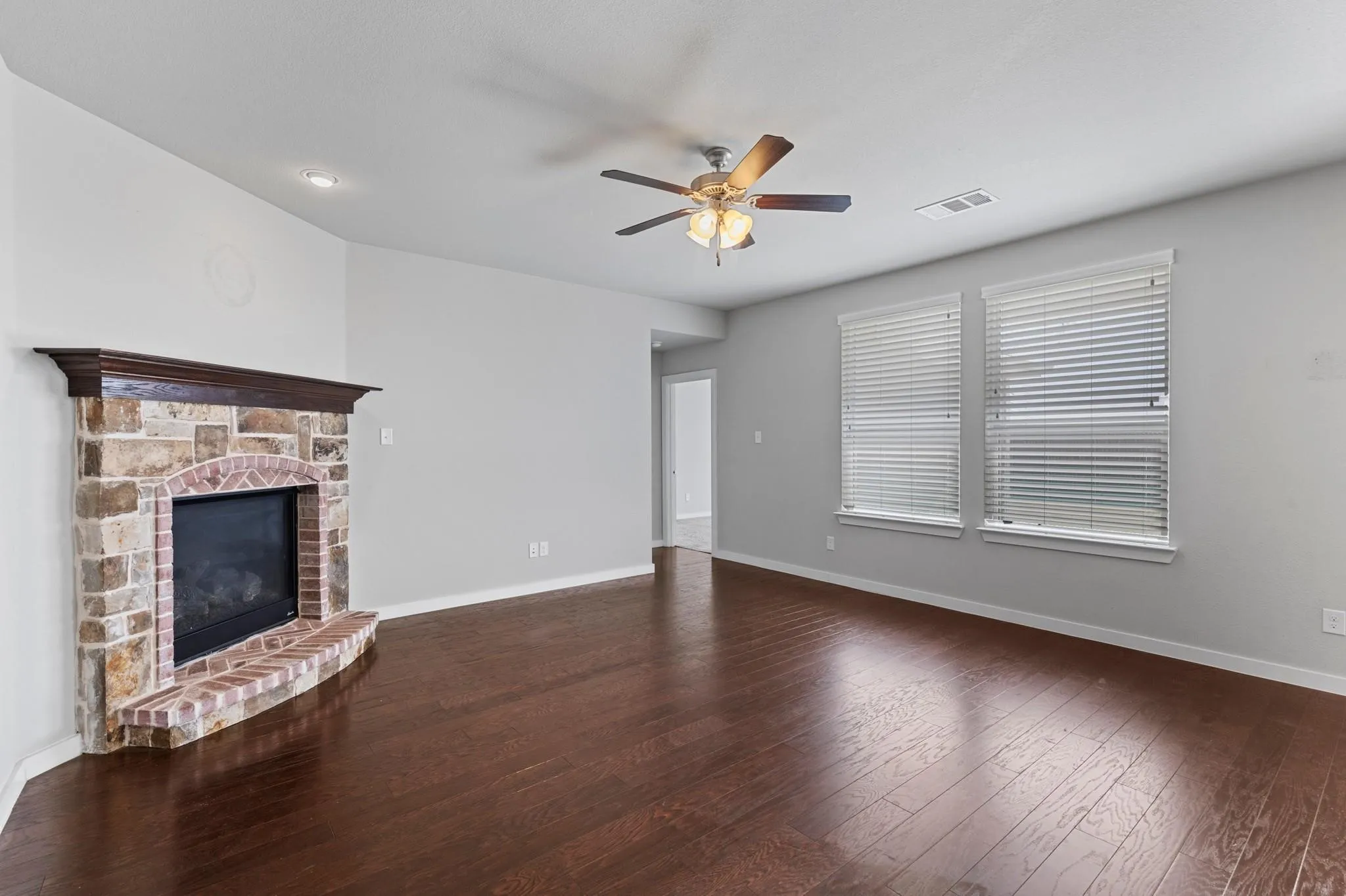 Living area featuring rich wood-finish flooring, a stone and brick fireplace with a wood mantel, two windows with blinds, recessed lighting, and a ceiling fan