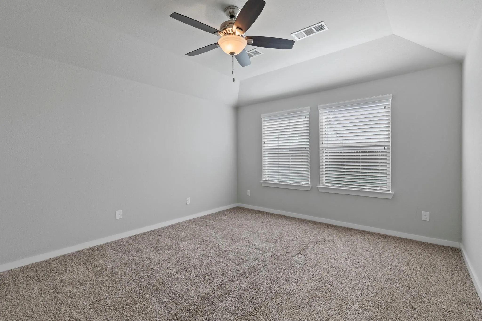 Carpeted interior space featuring a ceiling fan with integrated lighting, two windows with blinds, a vaulted ceiling, and light-colored walls