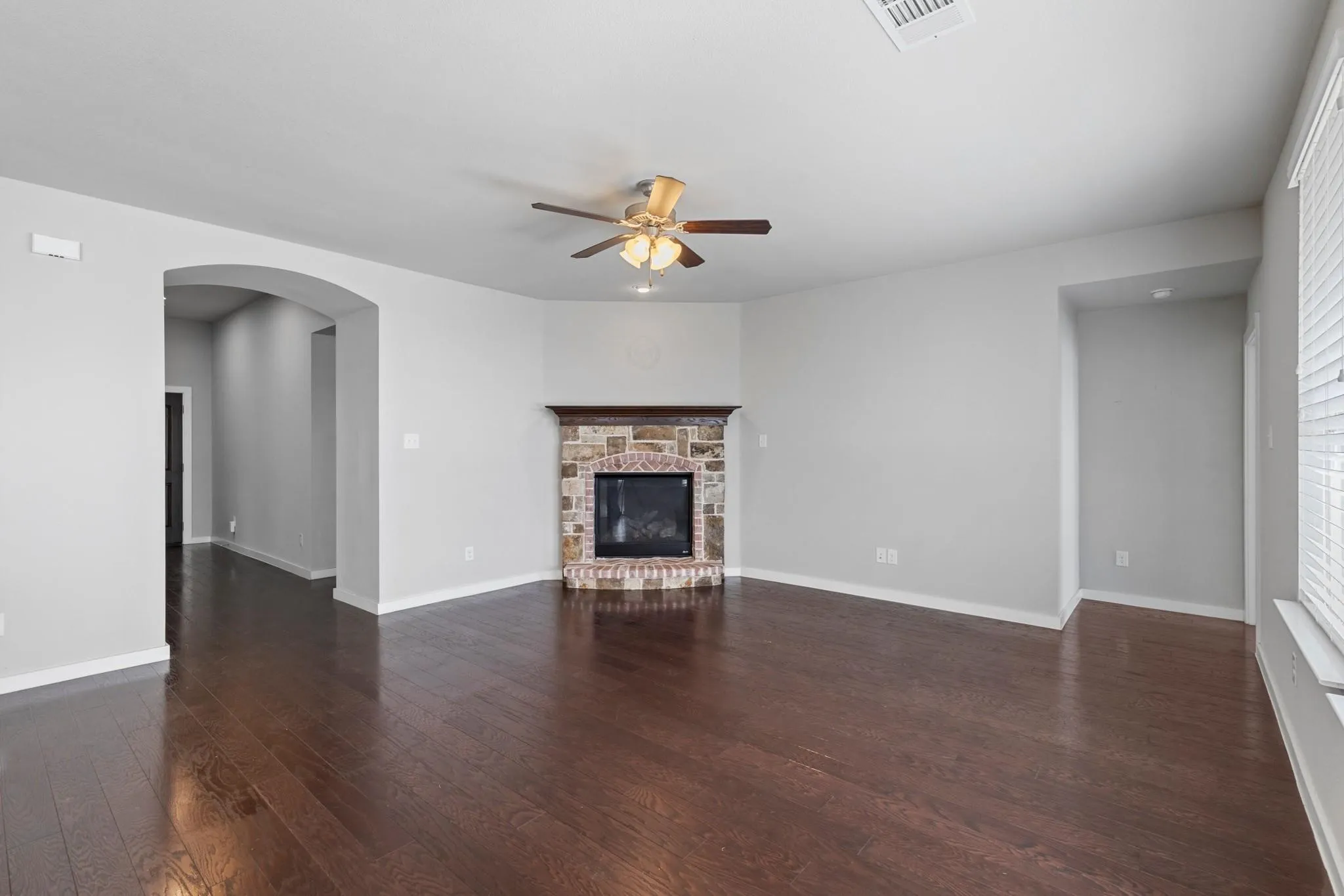 Spacious room featuring wood-finish flooring, a stone-faced corner fireplace with a wood mantel, and an arched entryway