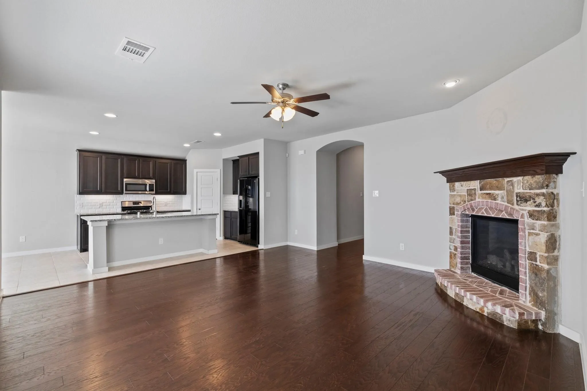 Open-concept living area featuring rich wood-finish flooring, a stone and brick fireplace with a wood mantle, and a kitchen island with seating