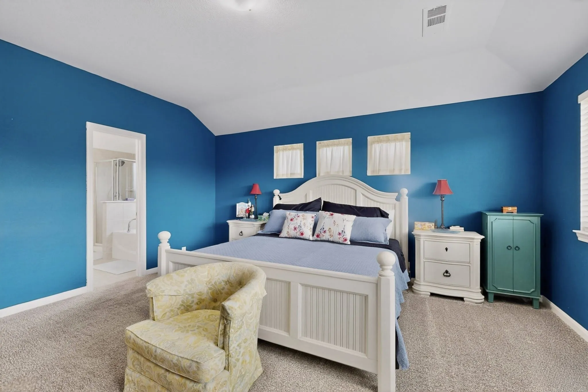 Carpeted bedroom featuring a vaulted ceiling, multiple windows with white blinds, and an entryway to a full bathroom with a shower and tub combination