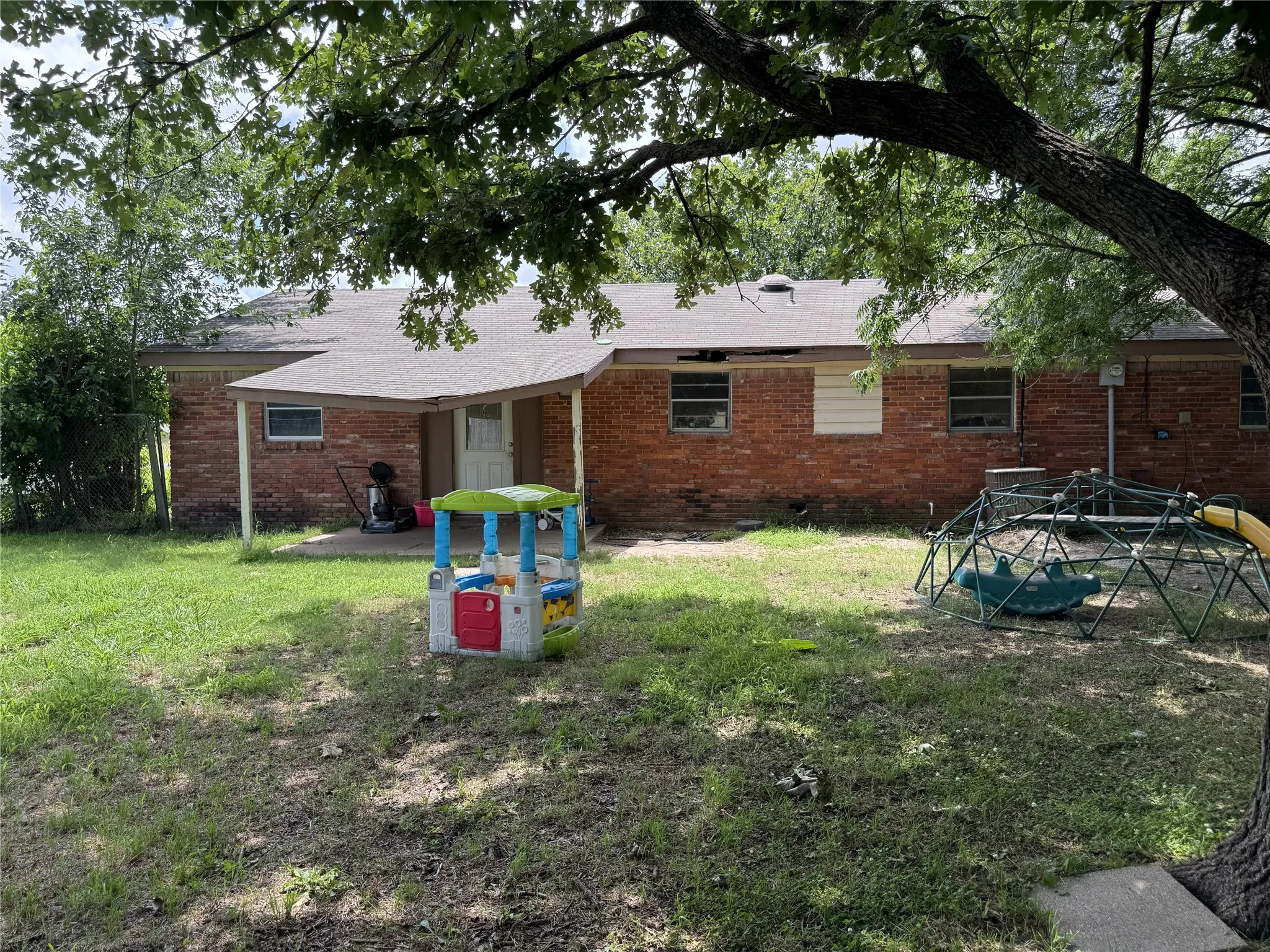 Expansive rear yard featuring a brick exterior home with multiple windows, a covered patio area, and mature trees providing shade