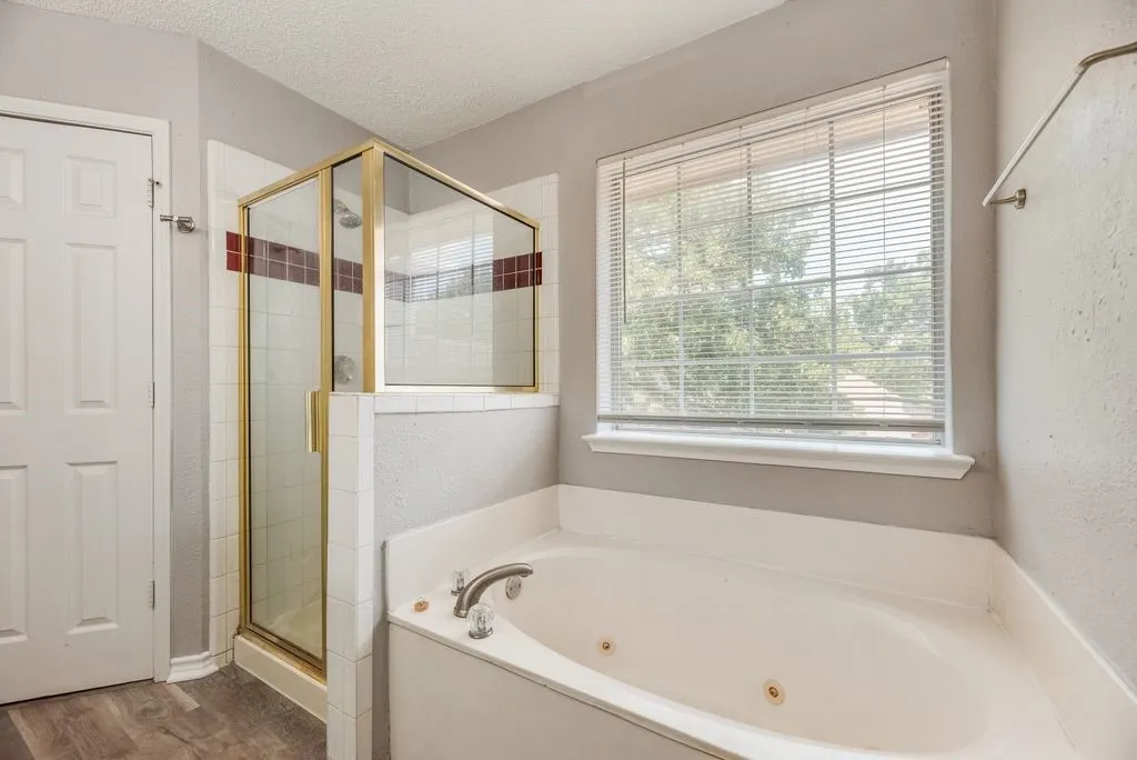 Spacious bathroom featuring a corner jetted soaking tub, separate glass-enclosed shower with tile surround, large window with blinds, and wood-finish flooring