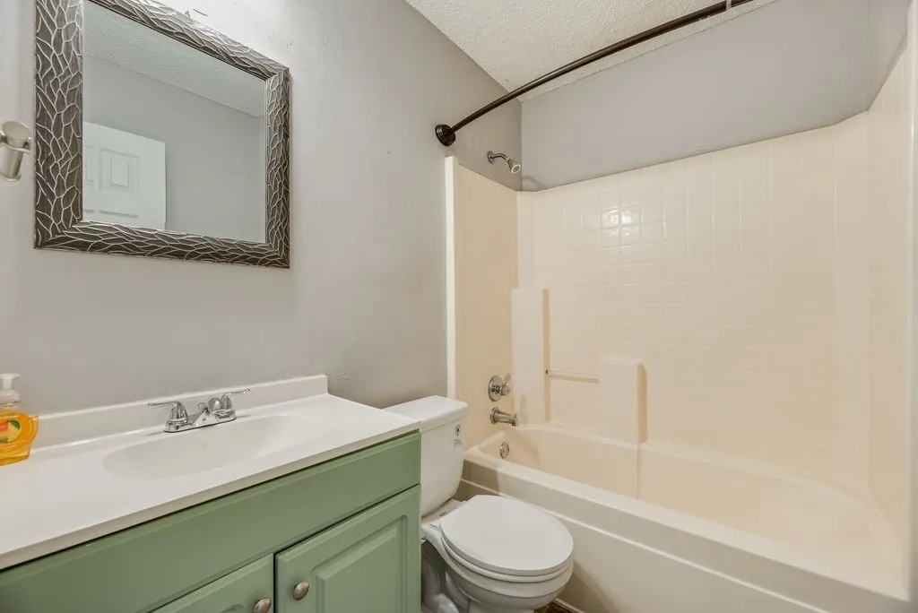 Bathroom featuring a tub/shower combination with tile surround, a vanity with a white countertop and integrated sink, a green-painted base cabinet with silver-tone hardware, and a framed mirror