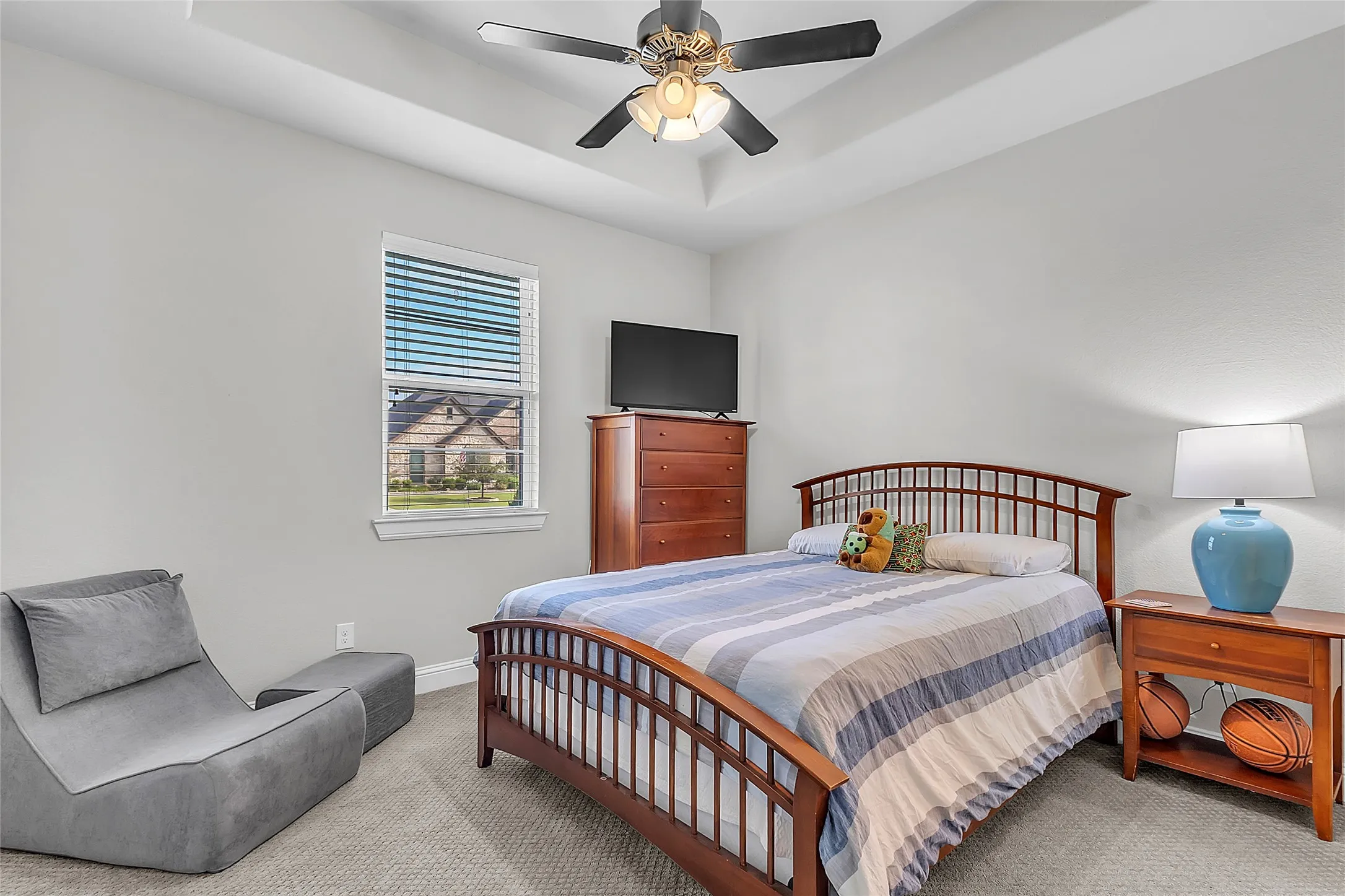 Carpeted secondary bedroom featuring a tray ceiling, ceiling fan with light, single window with blinds, and light gray wall color
