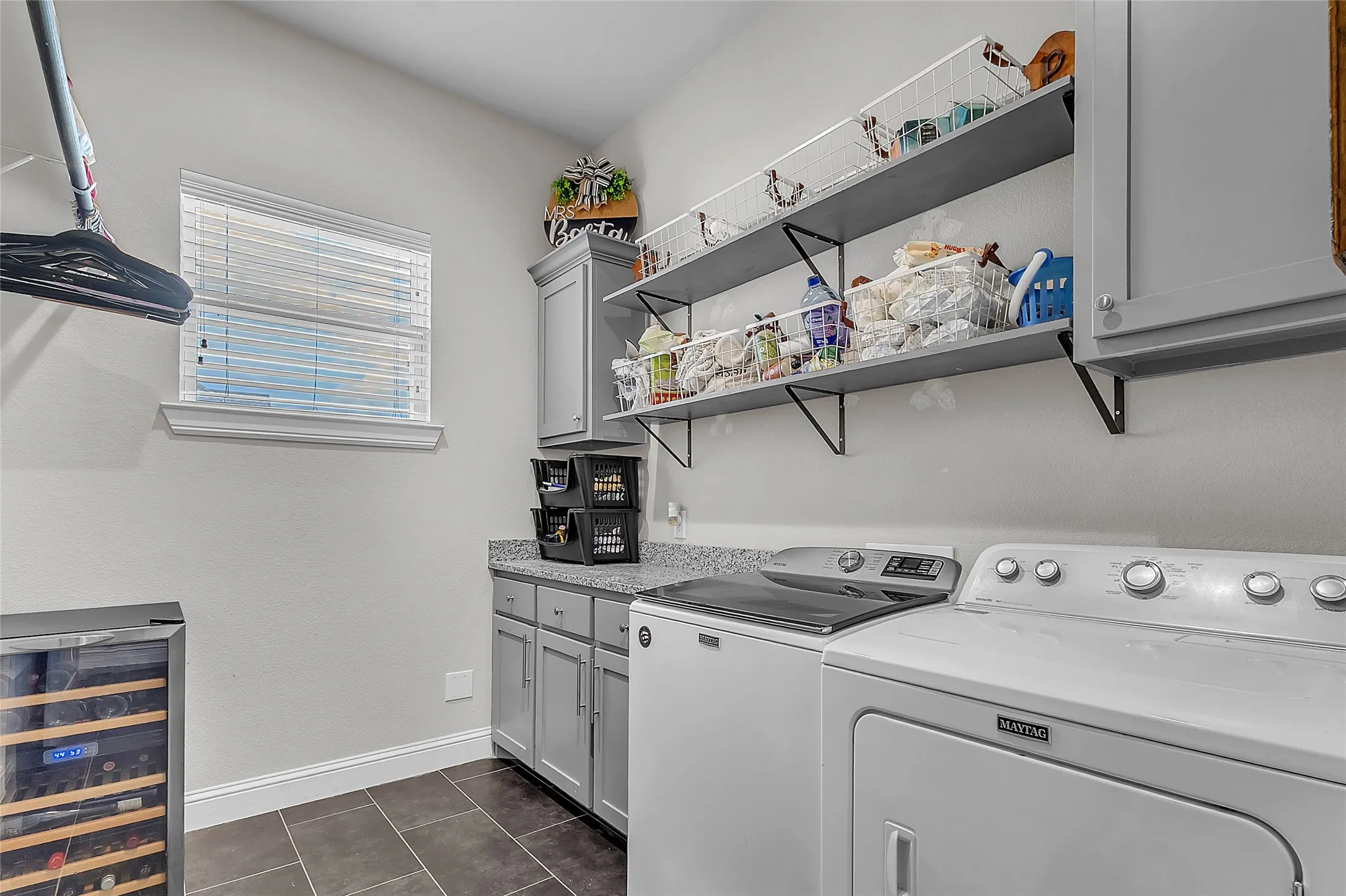 Dedicated laundry area featuring a window with blinds, cabinetry with a light-colored countertop, wall-mounted floating shelving, and dark tile flooring