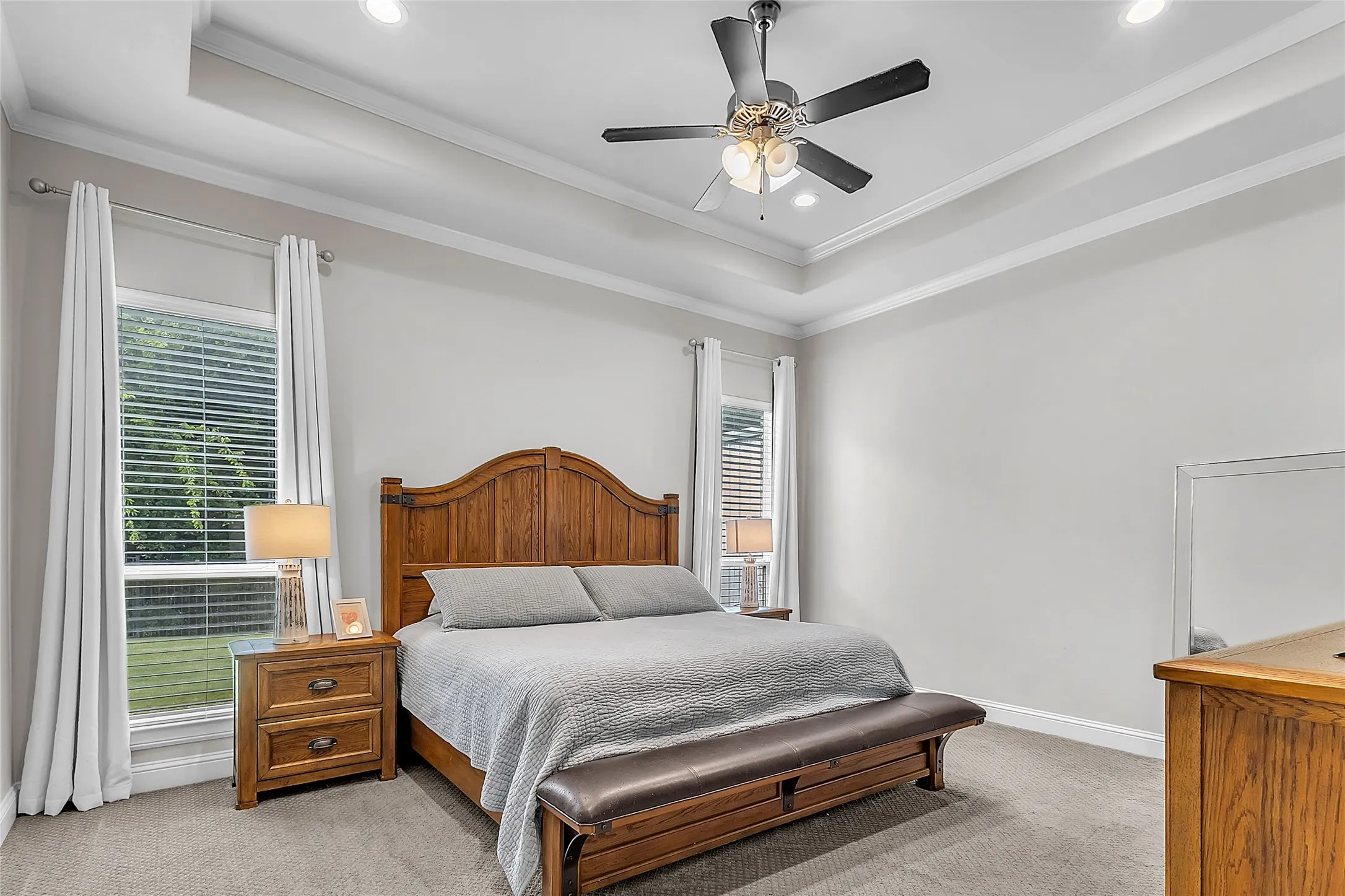 Primary bedroom with tray ceiling, crown molding, ceiling fan, two windows with tree-lined view of backyard, and carpeted flooring