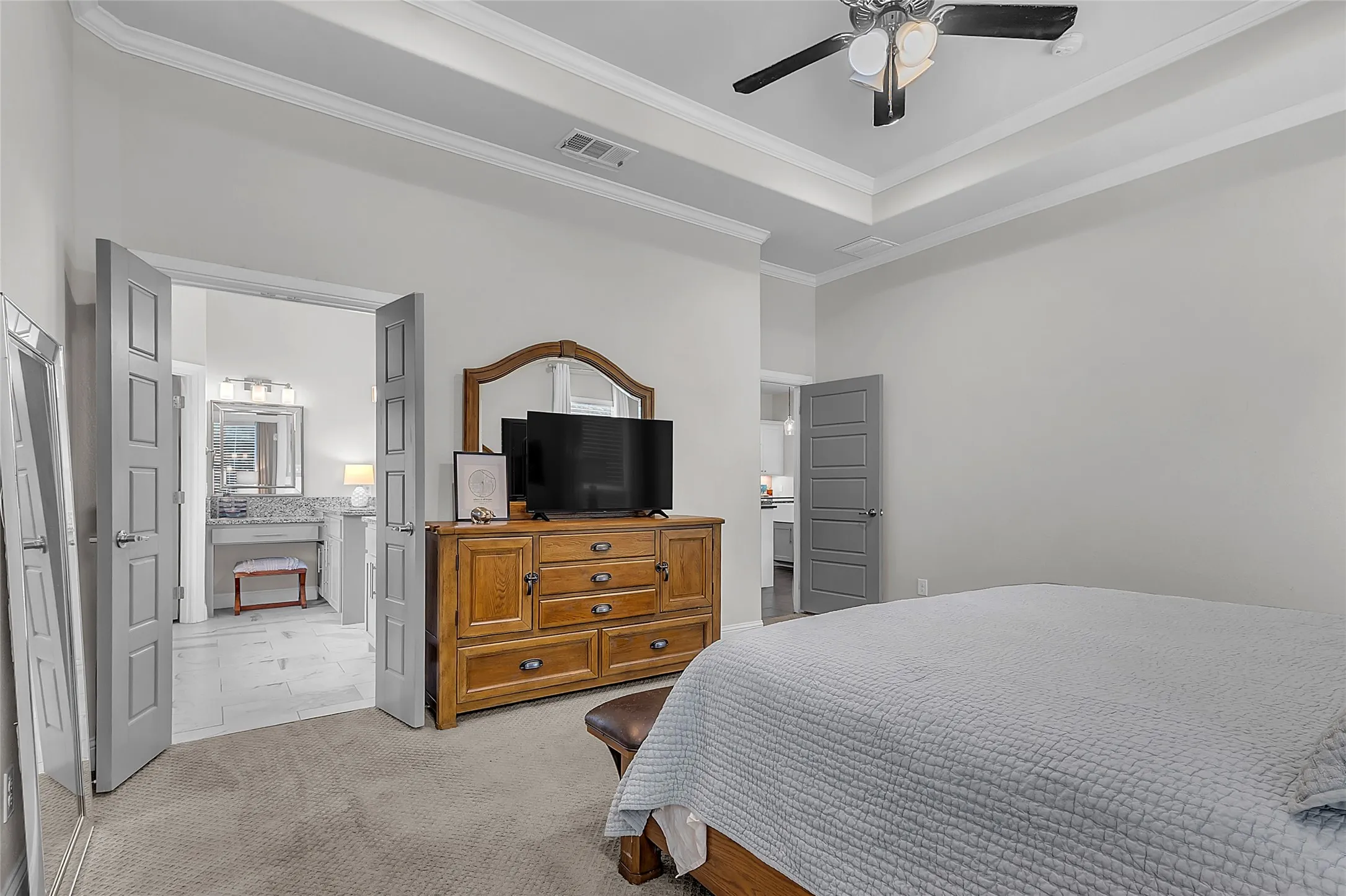 Primary bedroom with tray ceiling, crown molding, ceiling fan, two windows with tree-lined view of backyard, and carpeted flooring