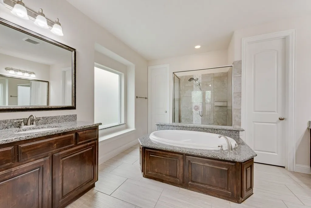 Bathroom featuring a built-in soaking tub with a granite surround, a glass-enclosed shower with tile walls, and a dual-sink vanity with dark wood cabinetry and granite countertops