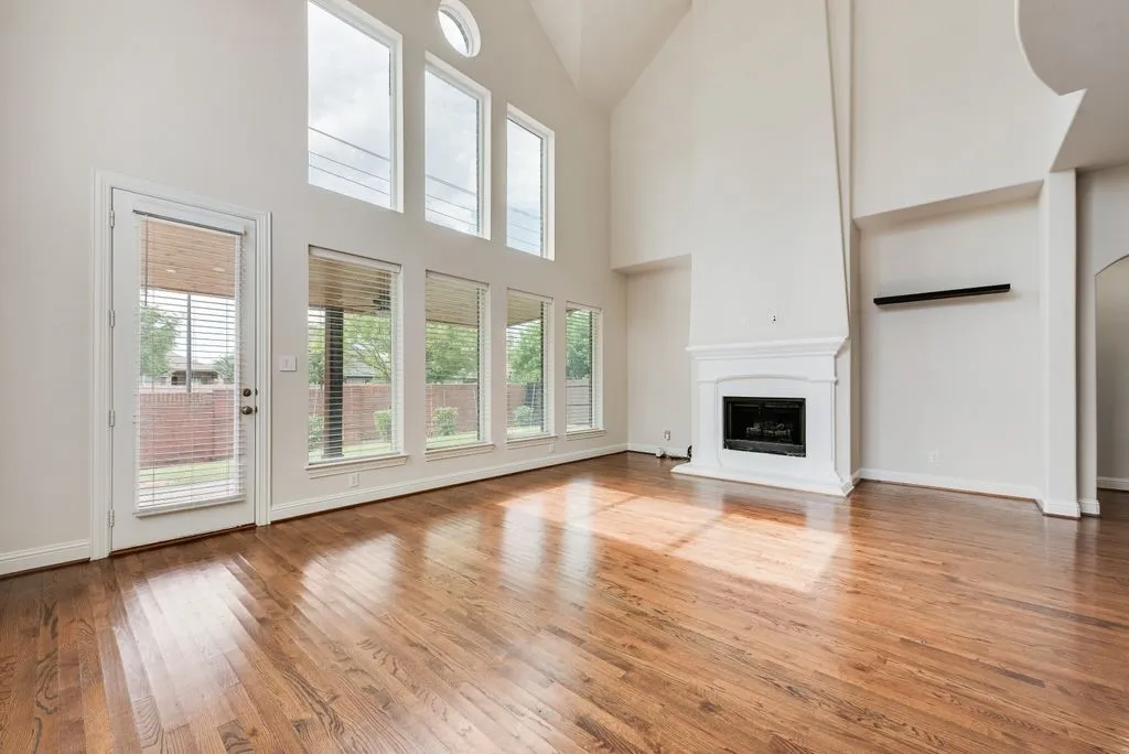 Spacious living area featuring high ceilings, wood-finish flooring, a white mantel fireplace, and multiple tall windows providing abundant illumination