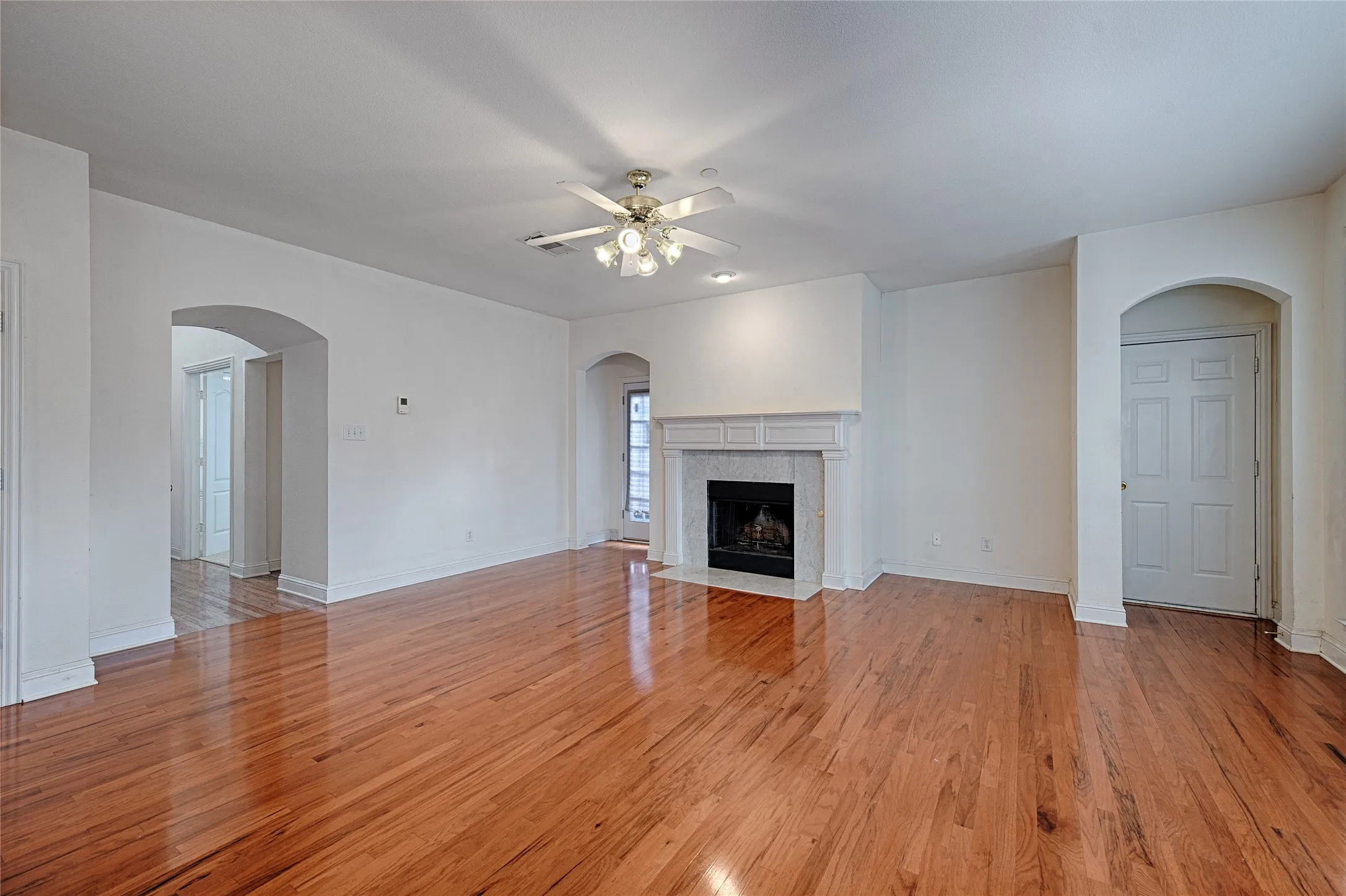 Spacious living area featuring rich wood-finish flooring, a central fireplace with a stone surround and decorative mantel, and a ceiling fan with integrated lighting