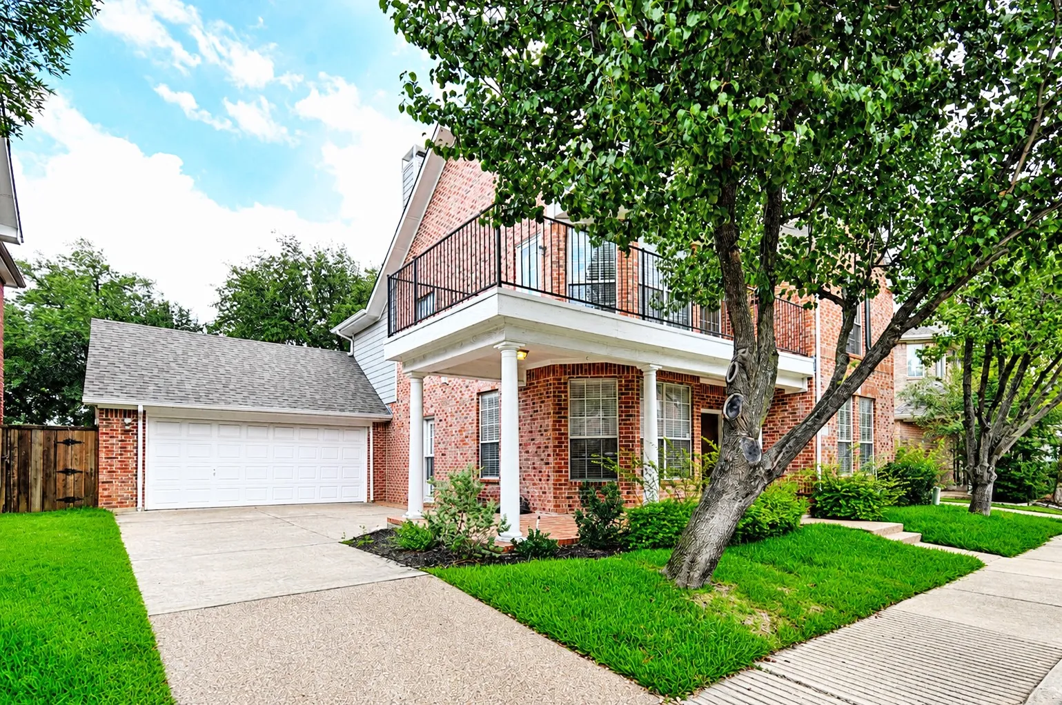 Brick exterior featuring a covered porch with white columns, an upper balcony with black iron railing, a two-car garage, and a concrete driveway