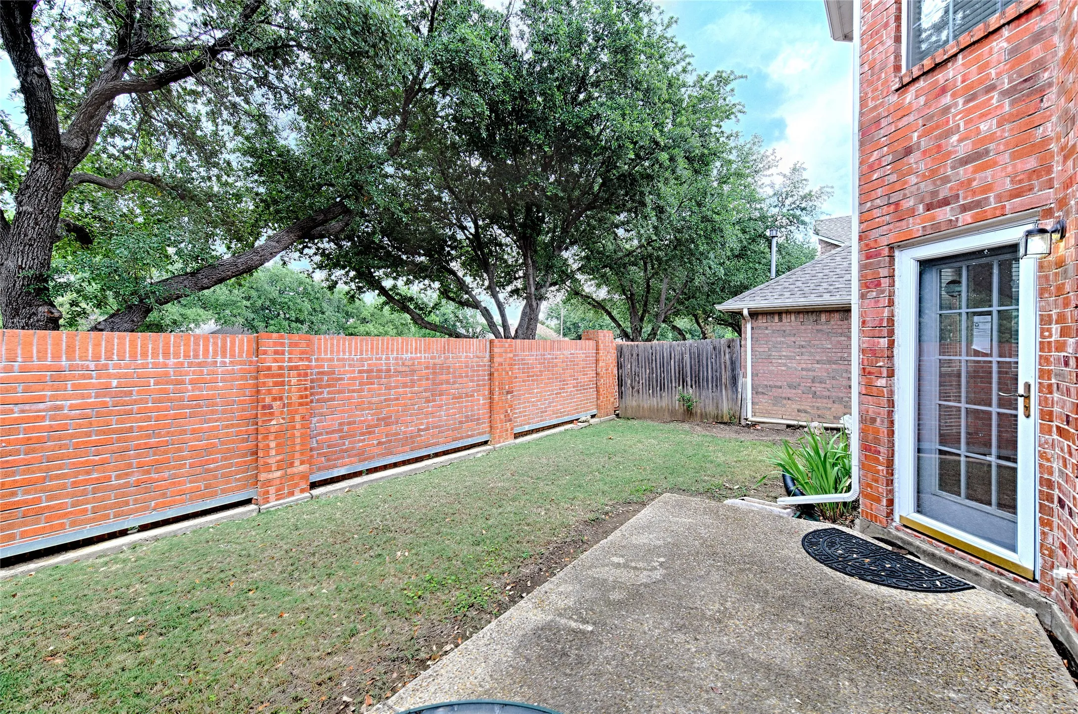 Private side yard featuring a red brick perimeter wall, mature trees, a concrete patio, and a screen door with gridded glass