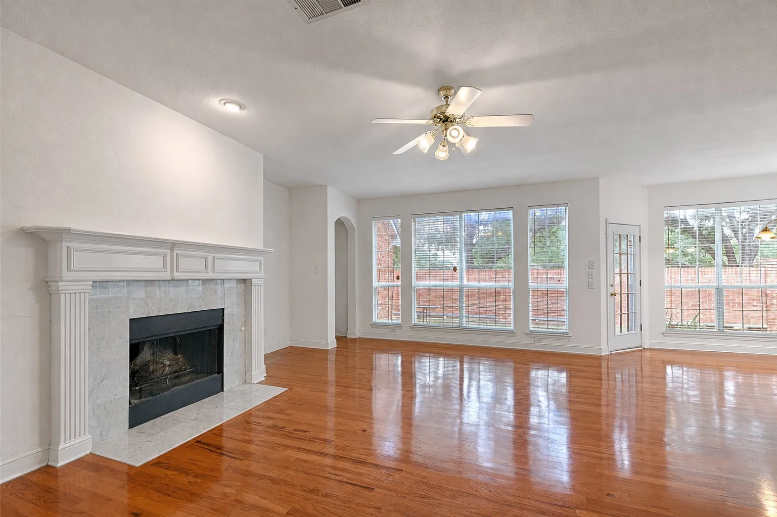 Spacious living area featuring a fireplace with a white mantel and light-toned tile surround, wood-finish flooring, a ceiling fan, and multiple large windows
