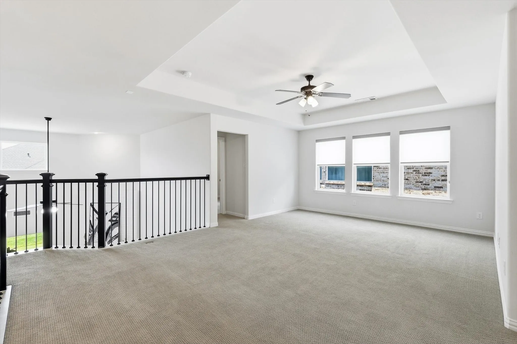 Spacious carpeted room featuring a tray ceiling, ceiling fan, and three windows with roller shades