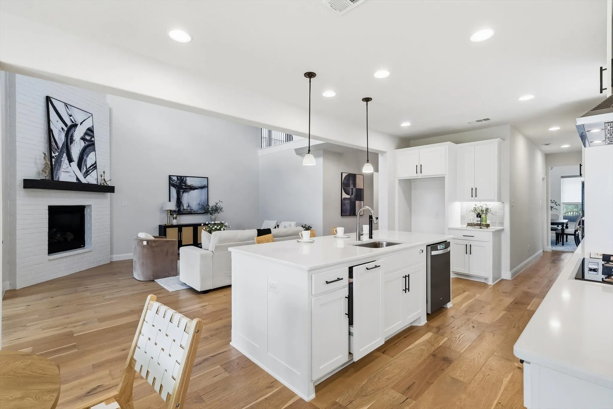 Open-concept living area featuring a large kitchen island with pendant lighting, white cabinetry with dark hardware, wood-finish flooring, and a white brick fireplace with a dark mantel