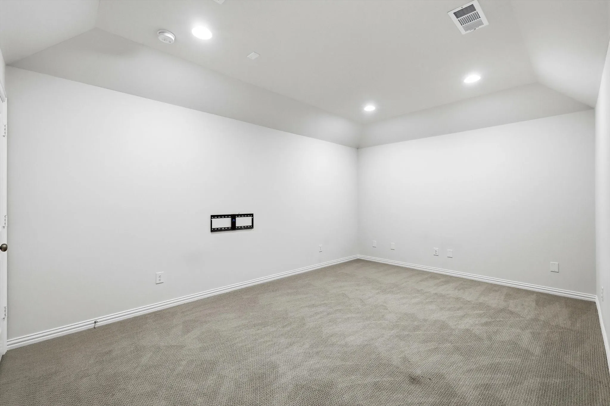 Spacious room featuring vaulted ceilings with recessed lighting, light-colored carpet flooring, a wall-mounted bracket, and white baseboards