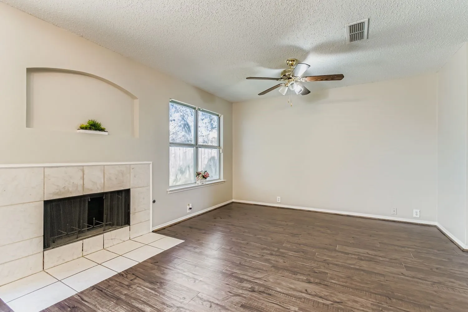 Living area featuring wood-finish flooring, a tiled fireplace with an arched niche, a multi-pane window, and a ceiling fan with integrated lighting