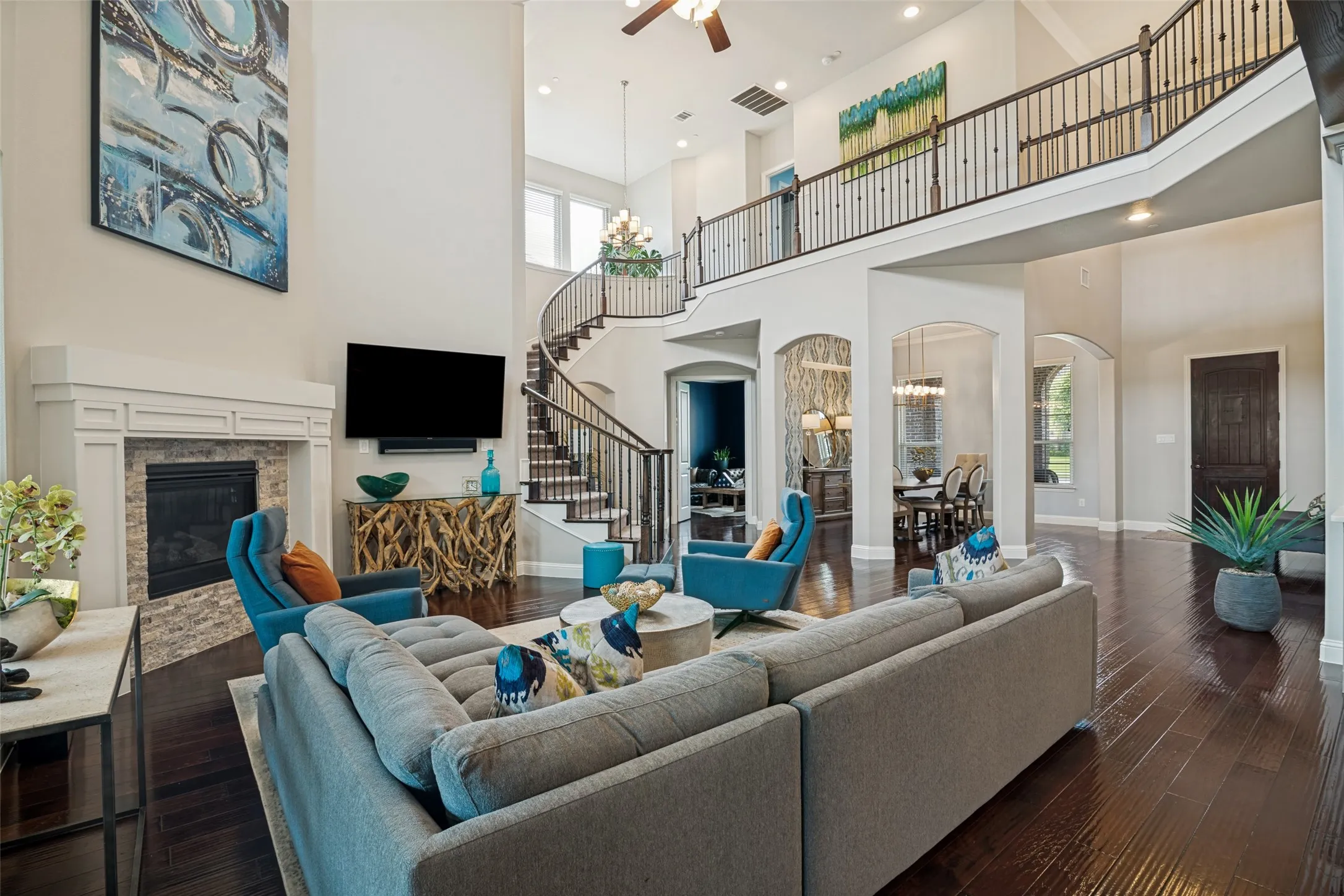 Two-story living area featuring dark pre engineered wood-finish flooring, a stone-faced fireplace with a white mantel, and a grand curved staircase with wrought iron balusters