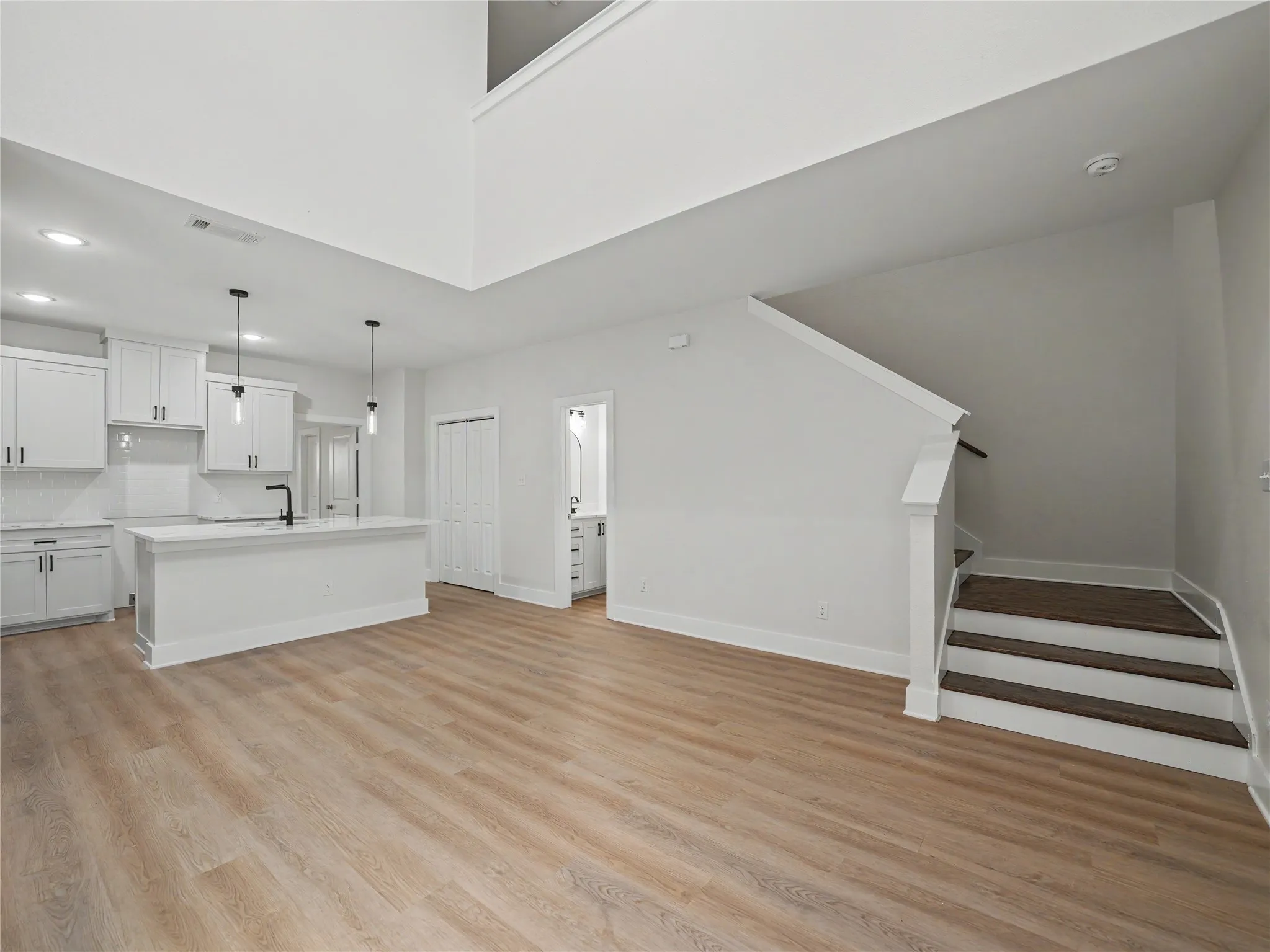Open-concept living space featuring wood-finish flooring, a contemporary kitchen with white cabinetry and island, matte black fixtures, and a staircase with dark wood treads