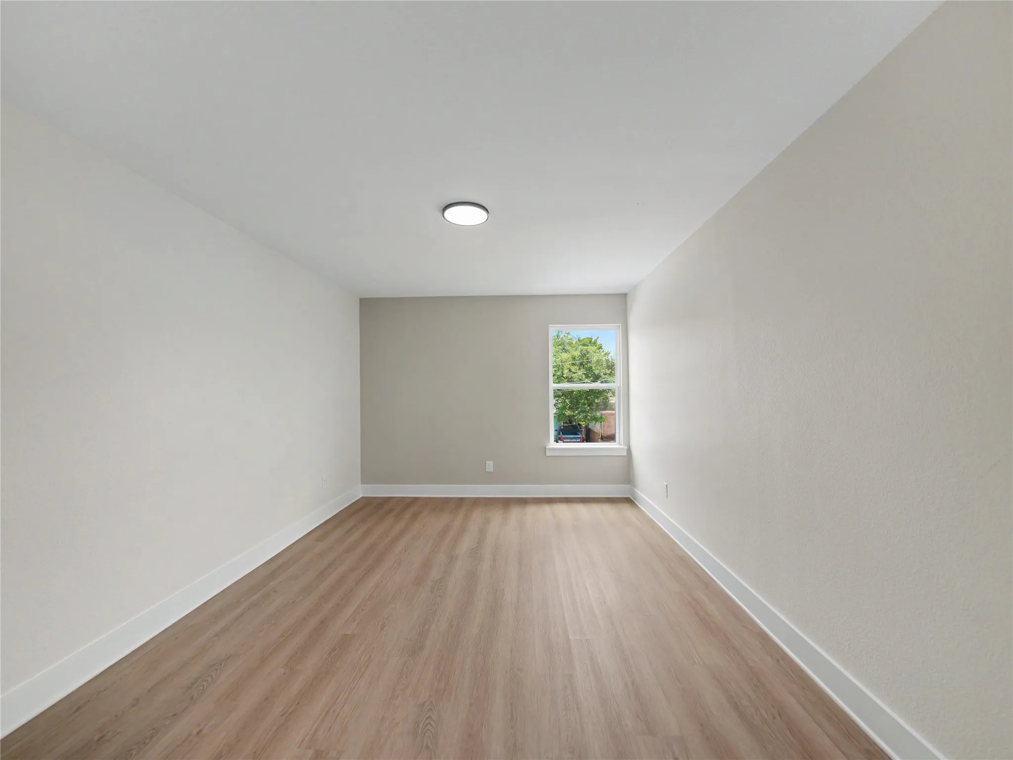Spacious room featuring wood-finish flooring, white baseboards, light neutral wall colors, a single window with white trim, and a flush-mount ceiling light fixture