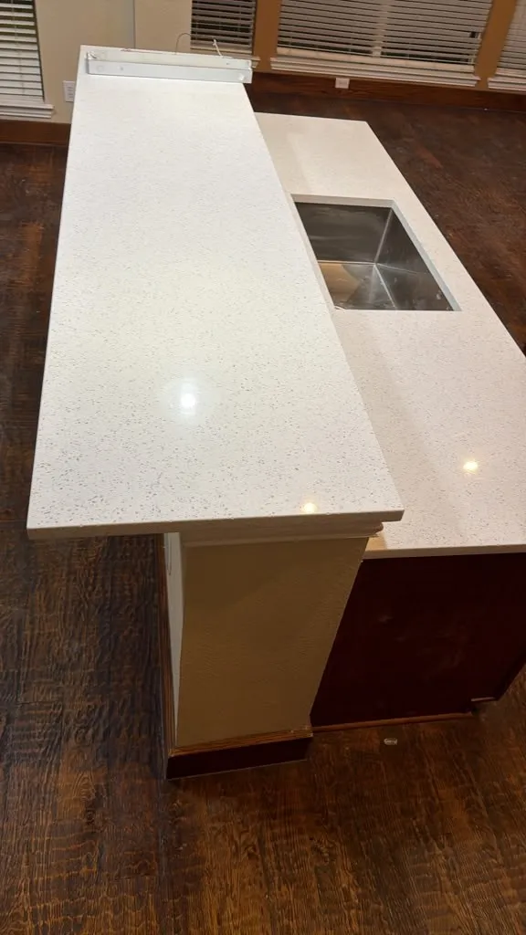 Kitchen island with white speckled countertop, integrated stainless steel sink, and wood-finish flooring