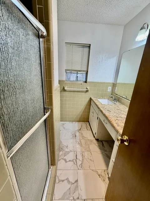 Shower enclosure with textured glass doors, light green tiled walls, a vanity with a granite-style countertop, a built-in sink, and patterned floor tiling